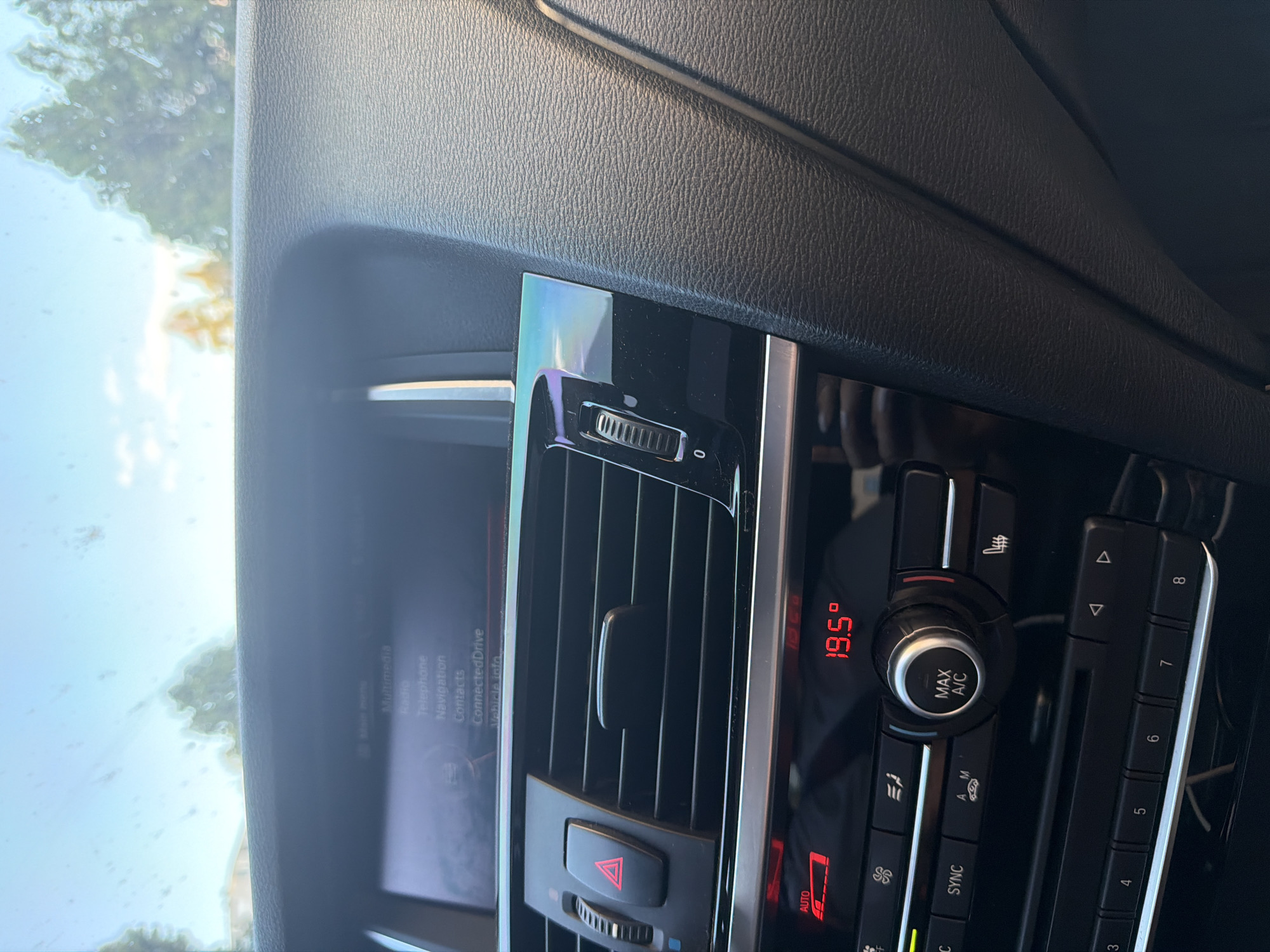 The image shows the interior of a car, focusing on the dashboard and center console area. Visible features include air vents, climate control buttons (including settings for temperature, fan speed, and air conditioning), an infotainment screen displaying menu options such as "Radio," "Telephone," "Navigation," and "ConnectedDrive," and several car-specific buttons arranged vertically. The interior is predominantly black, and the lighting suggests it is daytime.