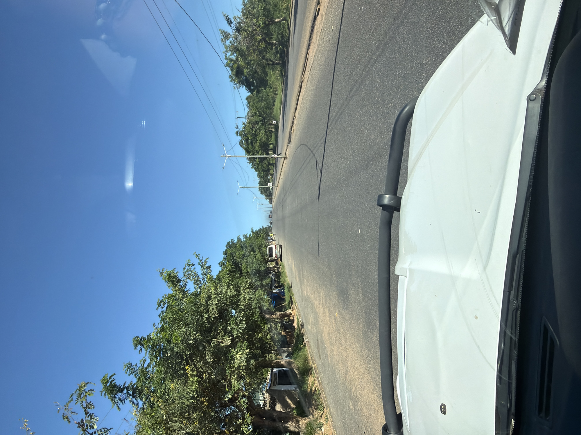 The image, taken from inside a vehicle, depicts a straight, asphalt road running through an urban area. The road is flanked by trees on both sides, some of which appear to be lining residential or commercial properties. Utility poles with visible wires run parallel to the road. The sky is clear and blue, with a visible reflection in the windshield. The vehicle's white exterior is visible in the foreground. Further down the road, several vehicles can be seen. The location is Unknown, Unknown.