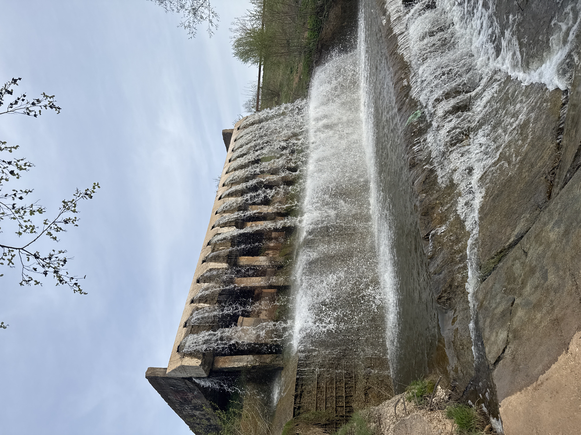 The image shows a cascading waterfall on a stepped concrete structure, likely a dam or spillway, at Kamennoye, Russia. Water flows in multiple streams down the terraced surface. Vegetation is visible on top of the structure. The sky is overcast.