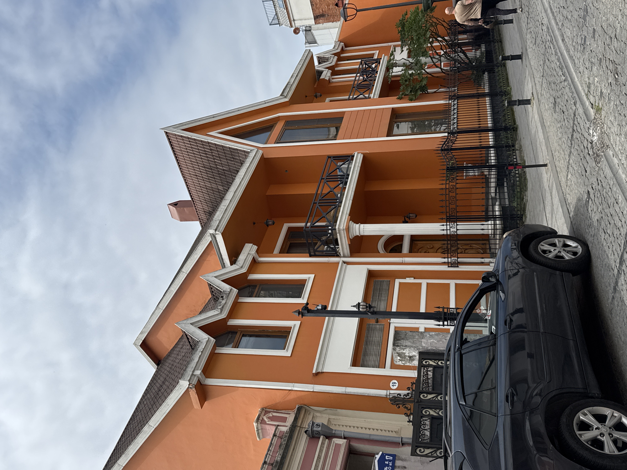 The image depicts a street scene in Batumi, Georgia, on a cloudy day. In the foreground, a dark grey SUV is parked on a cobblestone street. Behind the SUV, an eye-catching orange building with white trim dominates the scene. The building features multiple gabled roofs with brown tiles and decorative white trim along the eaves. There are several windows, some with balconies protected by black metal railings. A black metal fence separates the building's property from the sidewalk. Two streetlights are visible, one a black lamppost and another an orange decorative lamp. A man wearing a light brown t-shirt and dark pants walks away from the camera on the sidewalk. In the background, other buildings and a portion of a red-tiled roof are visible. A sign with Cyrillic text and the number "19" is affixed to the orange building.