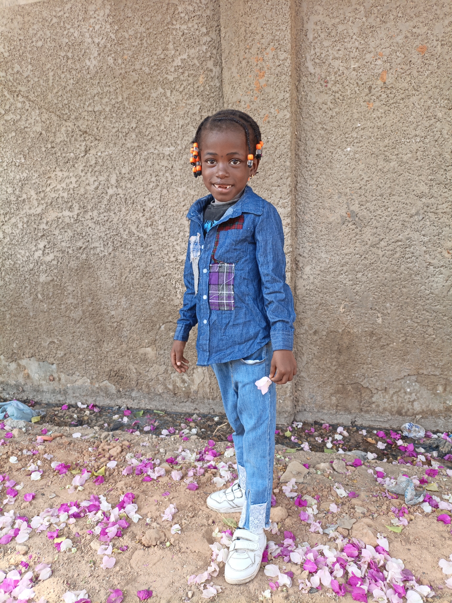A young child stands centered in front of a weathered concrete wall in Gawuna, Nigeria. The child, with beaded braids, wears a blue denim shirt with plaid patches and matching jeans, and white sneakers. The ground beneath is scattered with pink and white bougainvillea petals. A single flower is held in one hand. Debris, including plastic bags, is mixed with the petals. The background wall is textured, showing signs of age and wear.