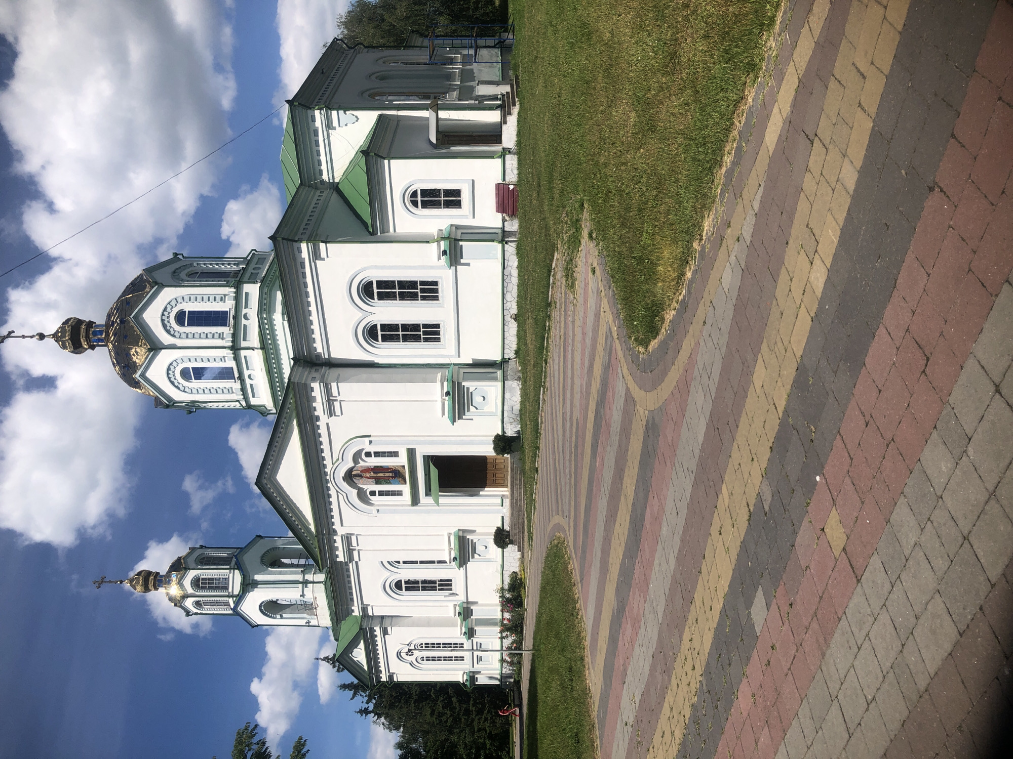 A white, orthodox church with a green roof and gold domes stands against a blue sky with scattered clouds. The church features multiple arched windows and a prominent bell tower. In front of the church is a paved walkway with multicolored bricks, curving around a patch of green grass. A person in a red outfit is visible walking on the grass to the left of the walkway. The environment appears to be a sunny day in a park-like setting.
