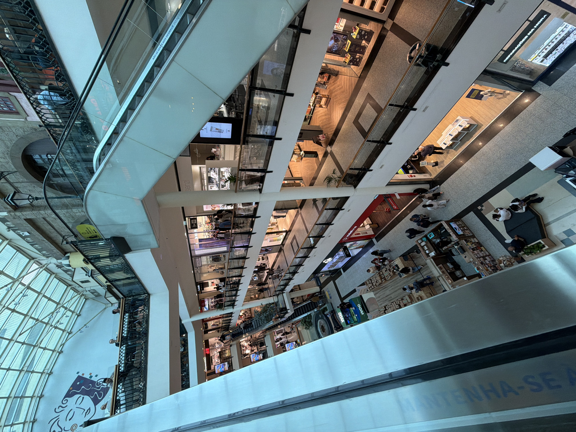 This is an interior shot of a multi-level shopping mall, possibly in Porto, Portugal, based on the local signage ("MANTENHA-SE" and "NOS DEGRAUS DO NOT"). The viewpoint is elevated, likely from an escalator or upper level, providing an overview of several floors. The architecture includes glass facades, escalators, and multiple retail spaces. The mall houses a variety of stores, including "Vodafone". People can be seen browsing and interacting within the shops on different floors. The overall atmosphere is that of a busy retail environment.