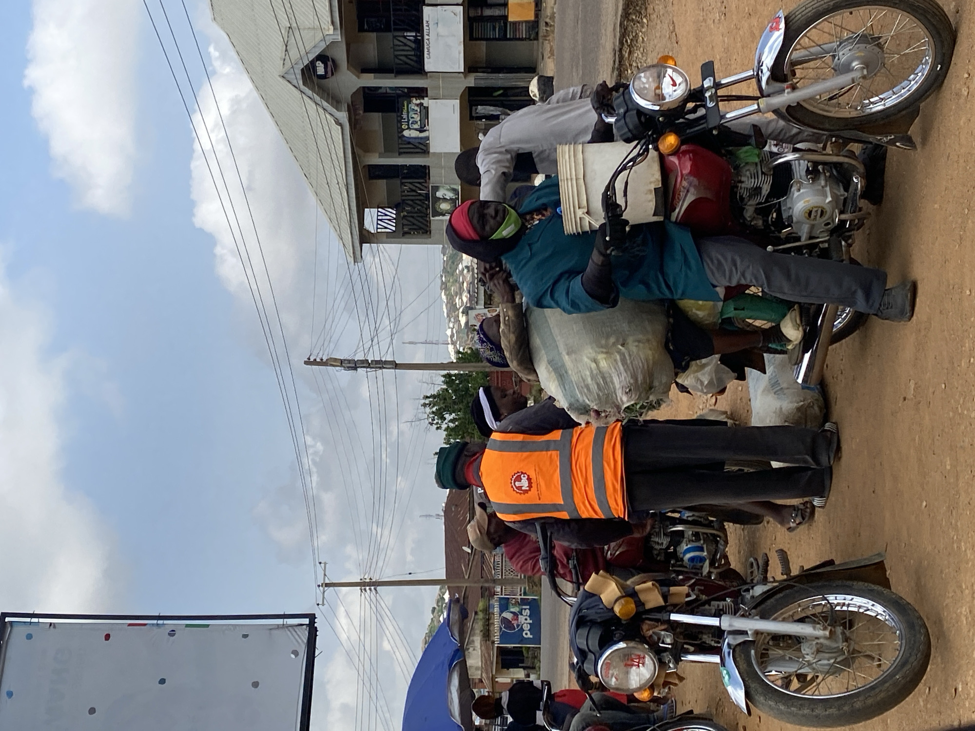 In Gwong, Nigeria, a group of individuals are gathered near a street, with several motorcycles present. One person, wearing an orange high-visibility vest, is standing and appears to be interacting with others. Another person is seated on a red motorcycle, holding a white bucket. Large, wrapped items are visible on the motorcycles, suggesting goods being transported. In the background, buildings and power lines are present, indicating an urban or semi-urban setting. The ground is dirt.