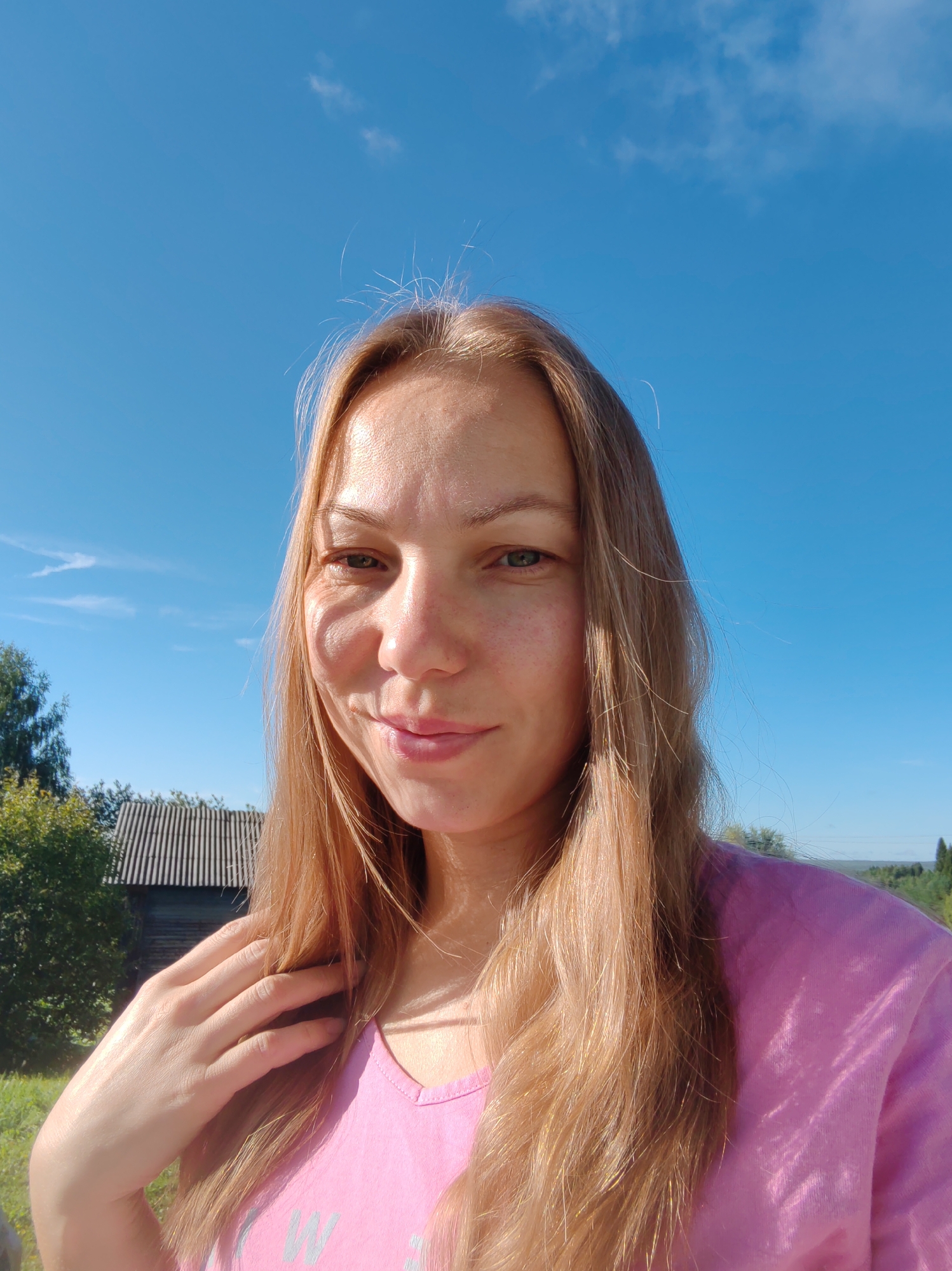 A young woman with long blonde hair is taking a selfie outdoors on a sunny day. She is wearing a pink t-shirt. Her left hand is touching her hair near her shoulder. In the background, there is a wooden building with a corrugated roof and green trees. The sky is bright blue with a few wispy clouds. The location is identified as Chukhlem, Russia.