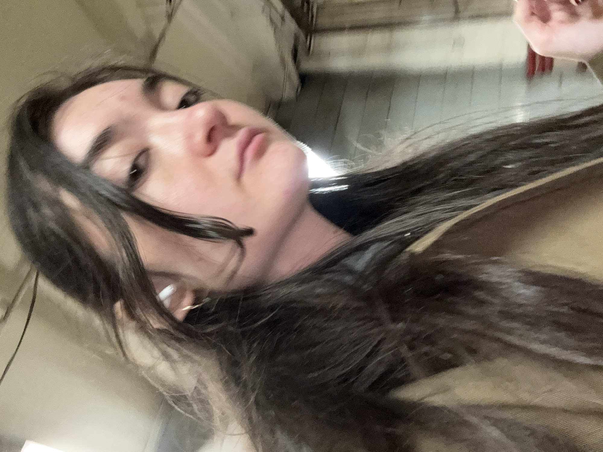 The image is a selfie of a young woman with dark hair, angled slightly upwards. She has fair skin and is wearing a white earbud in her left ear. Her lips are pursed, and her expression is somewhat neutral. Part of a brown jacket or coat is visible in the lower right corner. The background is blurry but appears to be an industrial or commercial space, possibly inside a building, with overhead structures and lighting. There is no indication of a specific location within New York City.