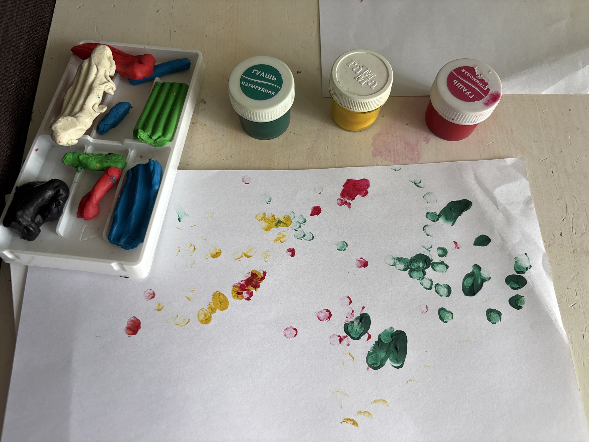 The image shows a craft or art activity. There's a white tray containing pieces of colored clay. Adjacent to the tray, there are three open jars of paint; one is green, one is yellow, and one is red. On a white piece of paper are several dots or fingerprints of the paints; some are red, some are yellow, and some are green. The word "гуашь" (gouache) is visible on the green and red paint jars, implying that the paint is gouache. The green jar also reads "изумрудная" (emerald) indicating the paint is emerald green. "Рубиновая" (Ruby) is visible on the red jar, identifying the paint as ruby red. "Izhevsk" is not visibly indicated or implied.