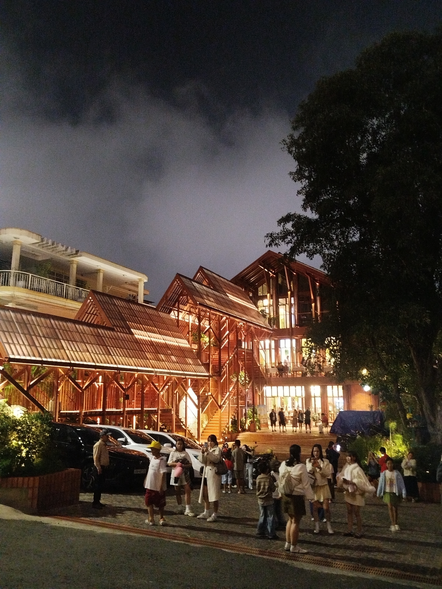 The image depicts an outdoor scene at night, possibly a gathering or a dining event, in Da Lat, Vietnam. The foreground shows several people standing and walking on a paved area, with cars parked behind them. The people are casually dressed, and some appear to be interacting or posing for photos. A young child in a yellow hat and red shorts stands out. In the background, a multi-story building with a distinctive wooden architecture and a tiled roof is brightly illuminated, suggesting it's a restaurant, resort, or public venue. The building has large windows revealing activity inside, and multiple levels with balconies and staircases. A large tree partially obscures the right side of the building, and the sky above is dark and cloudy, indicating it is nighttime. The overall atmosphere seems lively, with the warm lighting of the building creating a welcoming contrast to the dark sky. The location context suggests the scene is set in a temperate or mountainous region given Da Lat's climate. No visible text is present in the image.