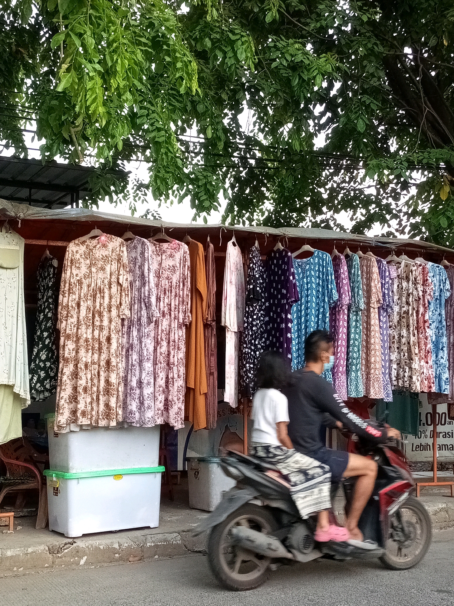 In a street market in Bekasi, Indonesia, two individuals are riding on a motorcycle. The man in front is wearing a black long-sleeved shirt, dark shorts, and a light blue face mask. The woman behind him is wearing a white short-sleeved shirt and a patterned skirt. They are moving from right to left across the frame. Behind them, a stall displays a variety of brightly colored dresses on hangers. The dresses feature floral and geometric patterns. In front of the stall, two large white plastic storage containers are stacked on top of each other, the top one with a green lid. The street is paved, and a concrete curb separates it from the market area. Lush green foliage from a large tree overhangs the scene, casting shadows. The overall impression is a snapshot of daily life in a bustling Indonesian marketplace.
