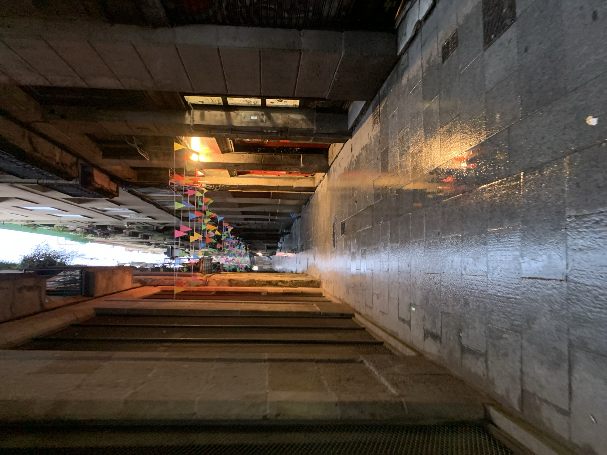 The image captures a view down a set of stone stairs in Barcelona, Spain, at night. The stairs are wide and lead down towards an area adorned with colorful triangular flags strung overhead. The walls alongside the stairs are made of stone and appear wet, reflecting light. Above, the underside of a bridge or building overhangs, creating a tunnel-like effect. A few lights illuminate the scene, casting reflections and shadows. There are no people visible in the immediate foreground.