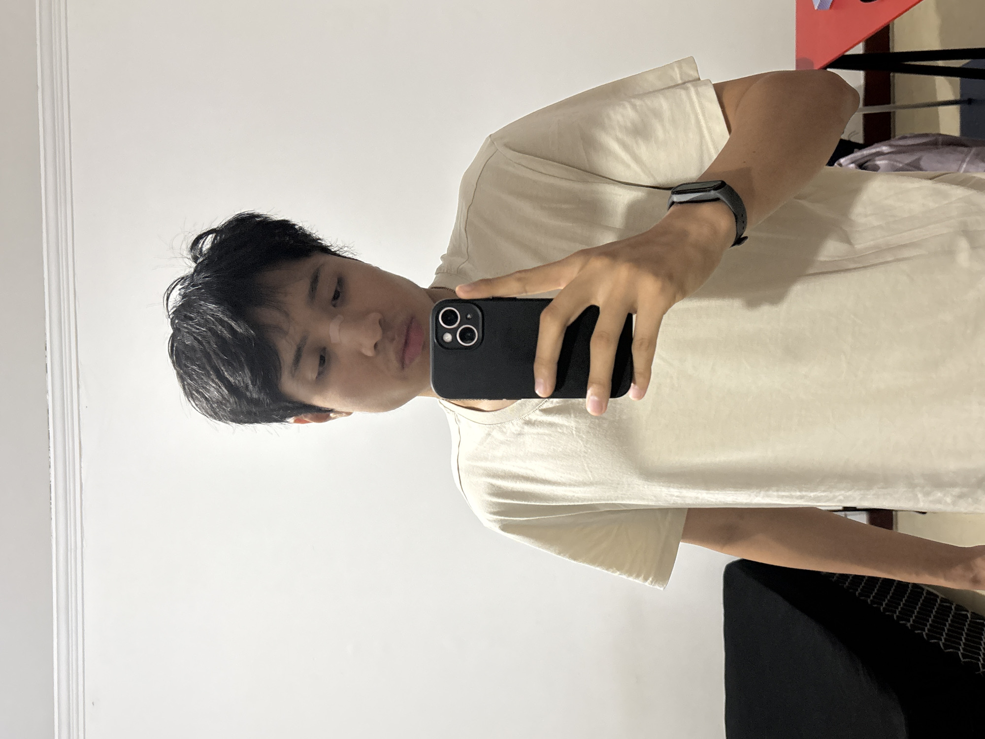 A person is taking a selfie in front of a white wall. The person is wearing a beige t-shirt and a black smart watch. They are holding a black smartphone in their right hand. The background includes a red table and black furniture. The location cannot be determined from the image.