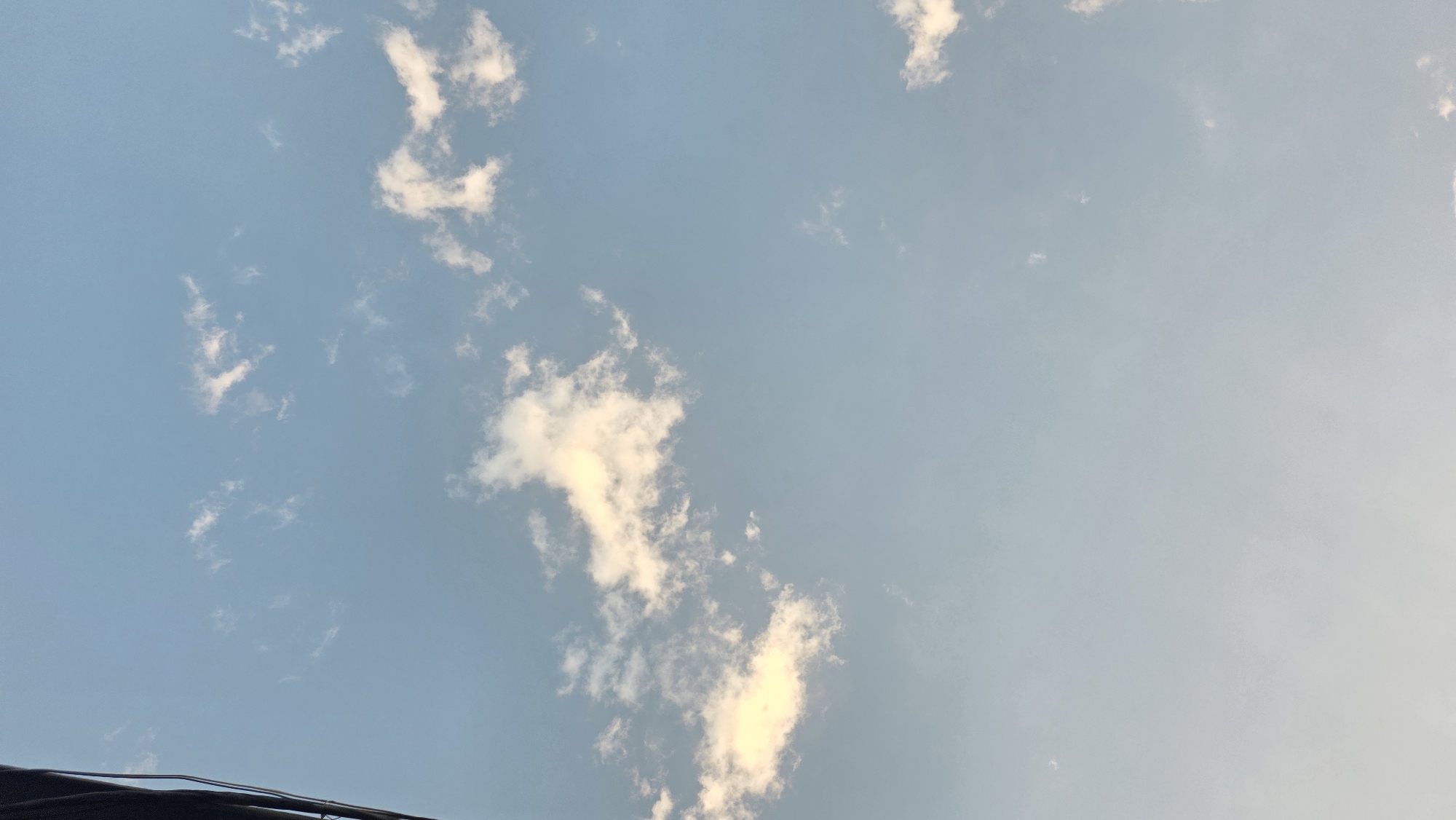 The image shows a pale blue sky with scattered wispy clouds, illuminated by soft sunlight. The lower part of the image transitions to a hazy, lighter shade of blue. On the far left edge, a dark, curved object is partially visible, suggesting the presence of a man-made structure, possibly a building or a utility pole, intruding into the natural scene. The overall impression is of a calm, open sky, characteristic of an outdoor environment in Chuy, Kyrgyzstan.