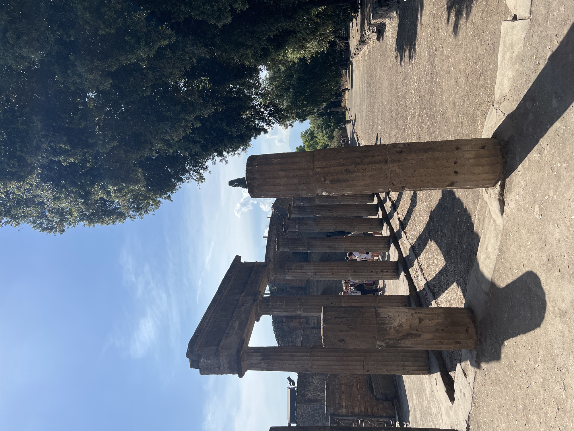 This image depicts a section of the ancient Roman city of Pompeii, Italy, under a clear blue sky with scattered clouds. The foreground features weathered stone columns, remnants of ancient structures, casting long shadows across a dusty pathway. In the background, more ruins are visible, including brick walls and archways, interspersed with lush green trees lining the path. Several tourists, dressed in casual summer attire, are present, walking along the path and observing the historical site. The atmosphere suggests a warm, sunny day, with the ruins preserved in a state of semi-restoration, allowing visitors to explore the remnants of this ancient civilization.