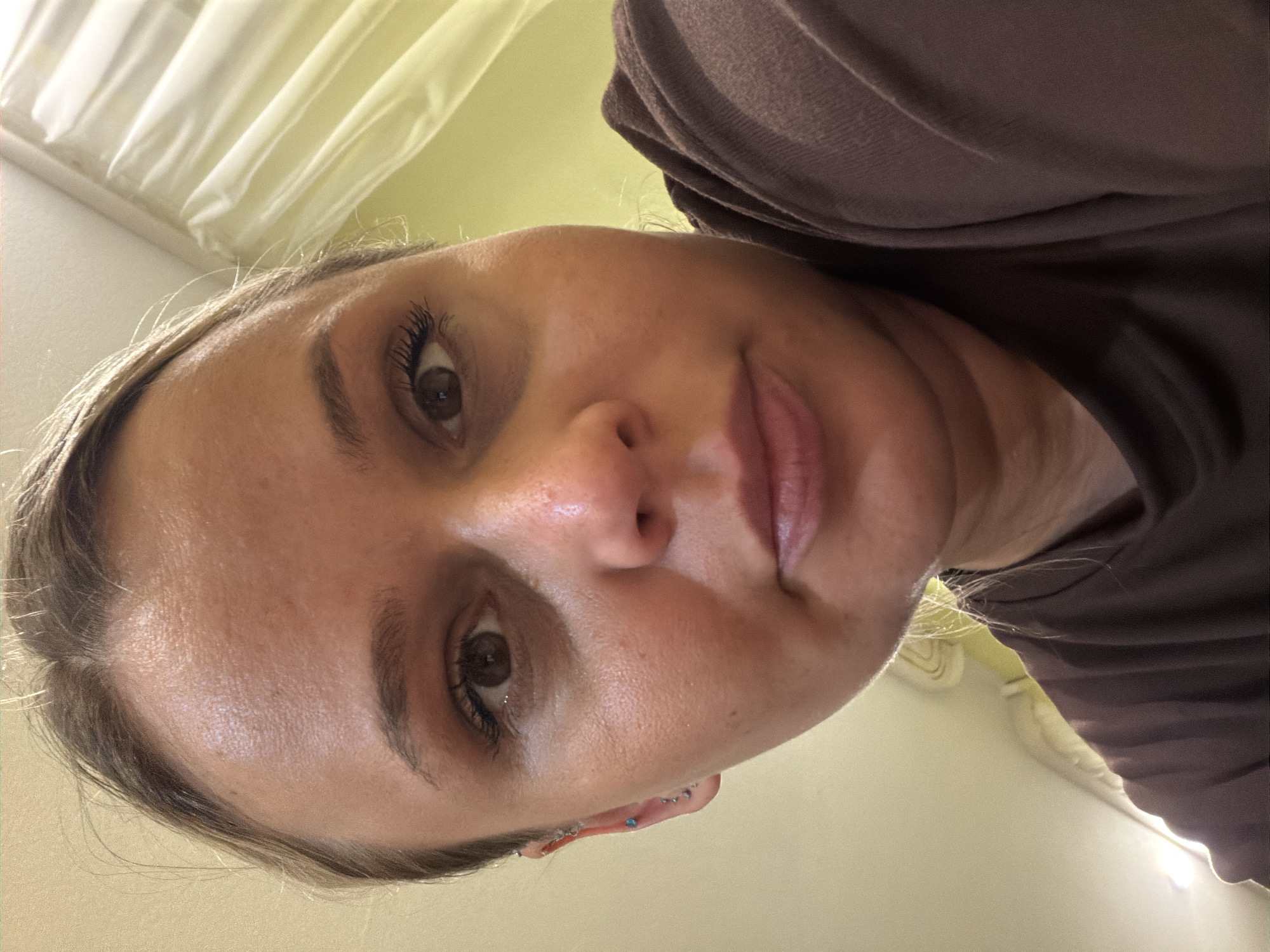 A close-up, angled self-portrait of a fair-skinned woman. Her face fills the frame, and she is wearing a brown top. Visible facial features include brown eyes, dark eyebrows, a light-colored nose, and pink lips. Her hair is light brown/blonde and pulled back. She has several ear piercings with blue jewelry on her left ear. The background appears to be an indoor setting with light-colored walls or curtains. No indication of a location within Sarapul, Russia, is present in the image.