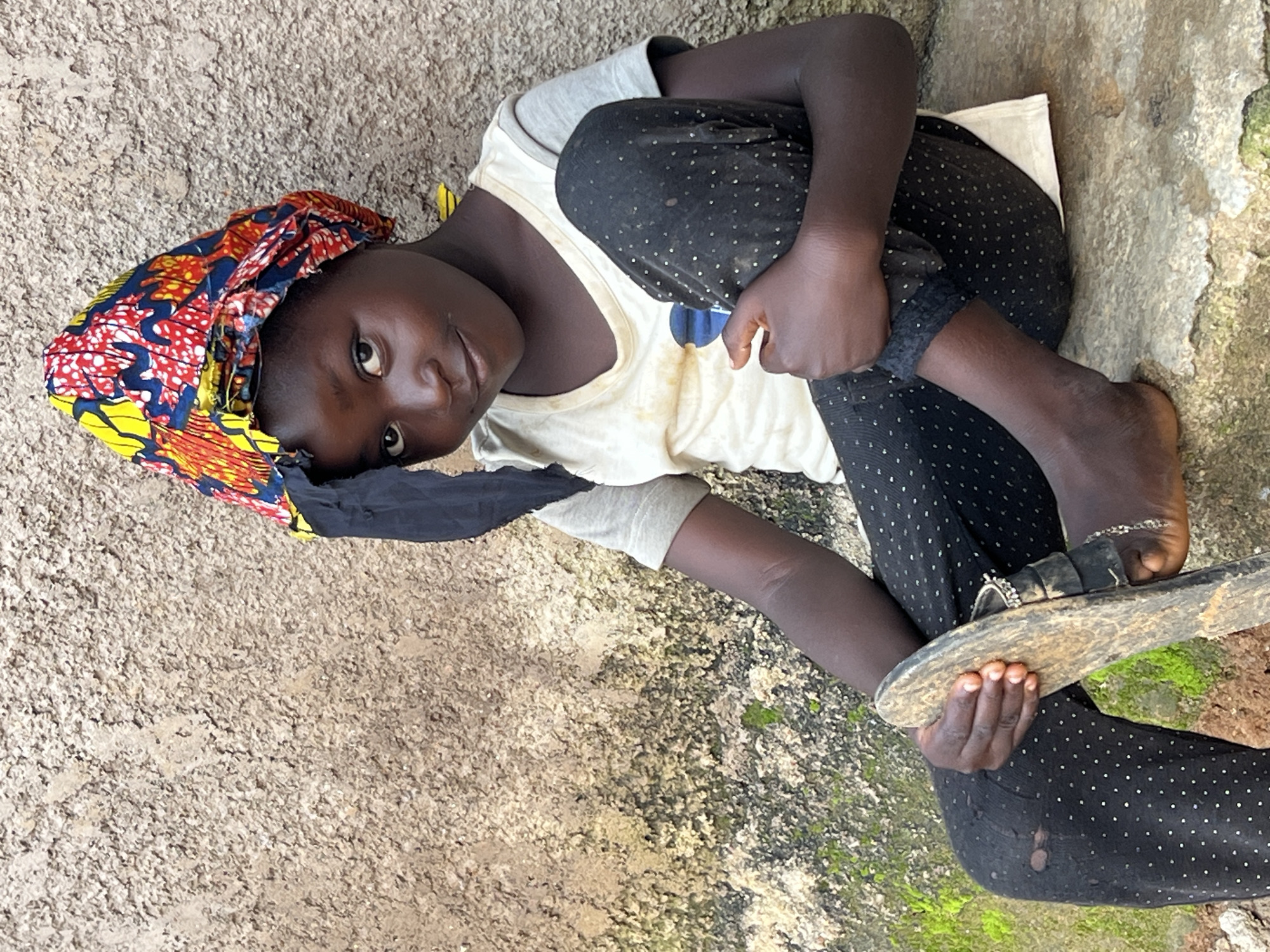 A young girl is sitting on a concrete ledge in front of a textured wall. She is wearing a colorful headscarf, a light-colored t-shirt, and black leggings with white polka dots. One leg is bent, and she is holding a sandal in her hands. Moss grows on the wall and the ledge. The background appears to be an outdoor setting, possibly in Jos, Nigeria.