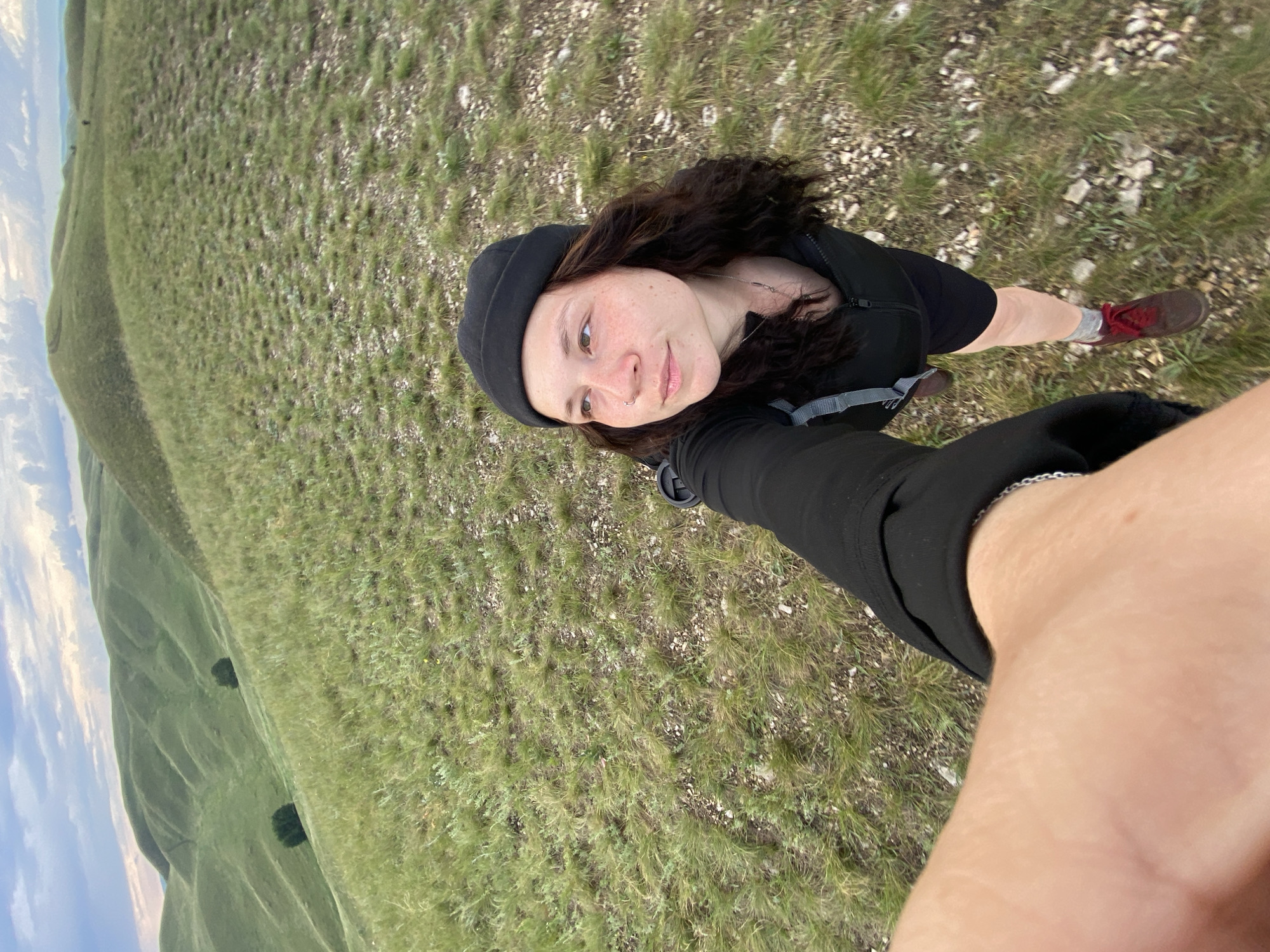 A person is taking a selfie in a vast, grassy, rolling landscape. The person, wearing a black beanie and black athletic clothing, is positioned in the foreground, looking slightly off-camera. The background features undulating green hills under a cloudy sky. The ground is covered in short, dry grass and scattered small stones. The overall scene suggests an outdoor activity like hiking in a rural or natural environment.