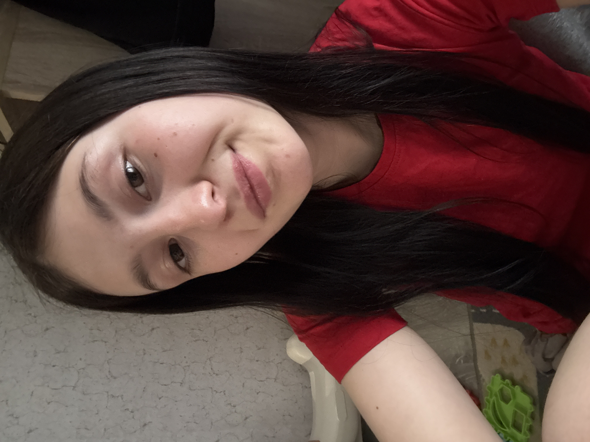 A portrait-oriented, slightly rotated image shows a young person, likely female, from the chest up, taking a close-up selfie. The subject has long, dark brown or black hair that falls over their shoulders and chest. They are wearing a plain red t-shirt. Their face is turned slightly to the right, and they have a soft, playful smirk or small smile on their lips. Their eyes are dark, and one eye is clearly visible, looking towards the camera. The skin appears fair with a few faint freckles or blemishes visible on the cheek.
The background on the left side of the image features a light grey, textured surface, possibly a blanket, carpet, or upholstery. On the right, behind the subject's head and body, a darker, patterned fabric or material is visible, which appears to be a blanket or rug. A small part of a white object, possibly a piece of furniture or an armrest, is also visible near the bottom left. Towards the bottom right corner, a distinctive bright green plastic object, possibly a toy or household item, is partially visible. The overall setting appears to be an indoor, casual environment. There is no visible text in the image.