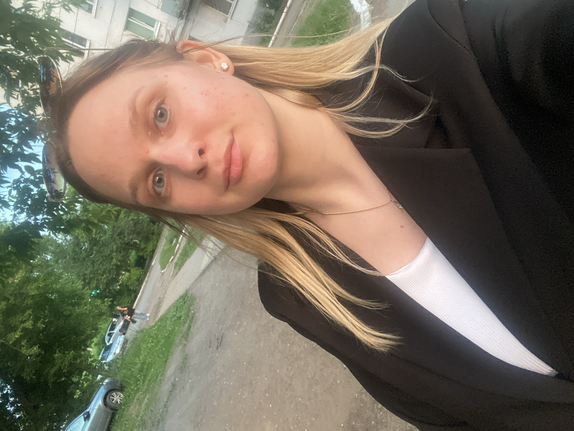 The image shows a young woman with blonde hair and blue eyes, taking a selfie outdoors. She is wearing a black blazer over a white top and a delicate gold necklace. Her sunglasses are resting on top of her head. The background suggests a residential area, with trees, a paved walkway, and parked cars. The presence of apartment buildings in the background, typical of Soviet-era architecture, aligns with the visual cues of Chaikovsky, Russia, a city known for its industrial and residential sectors. The lighting indicates it is likely late afternoon or early evening. The woman appears to be alone in the immediate foreground.