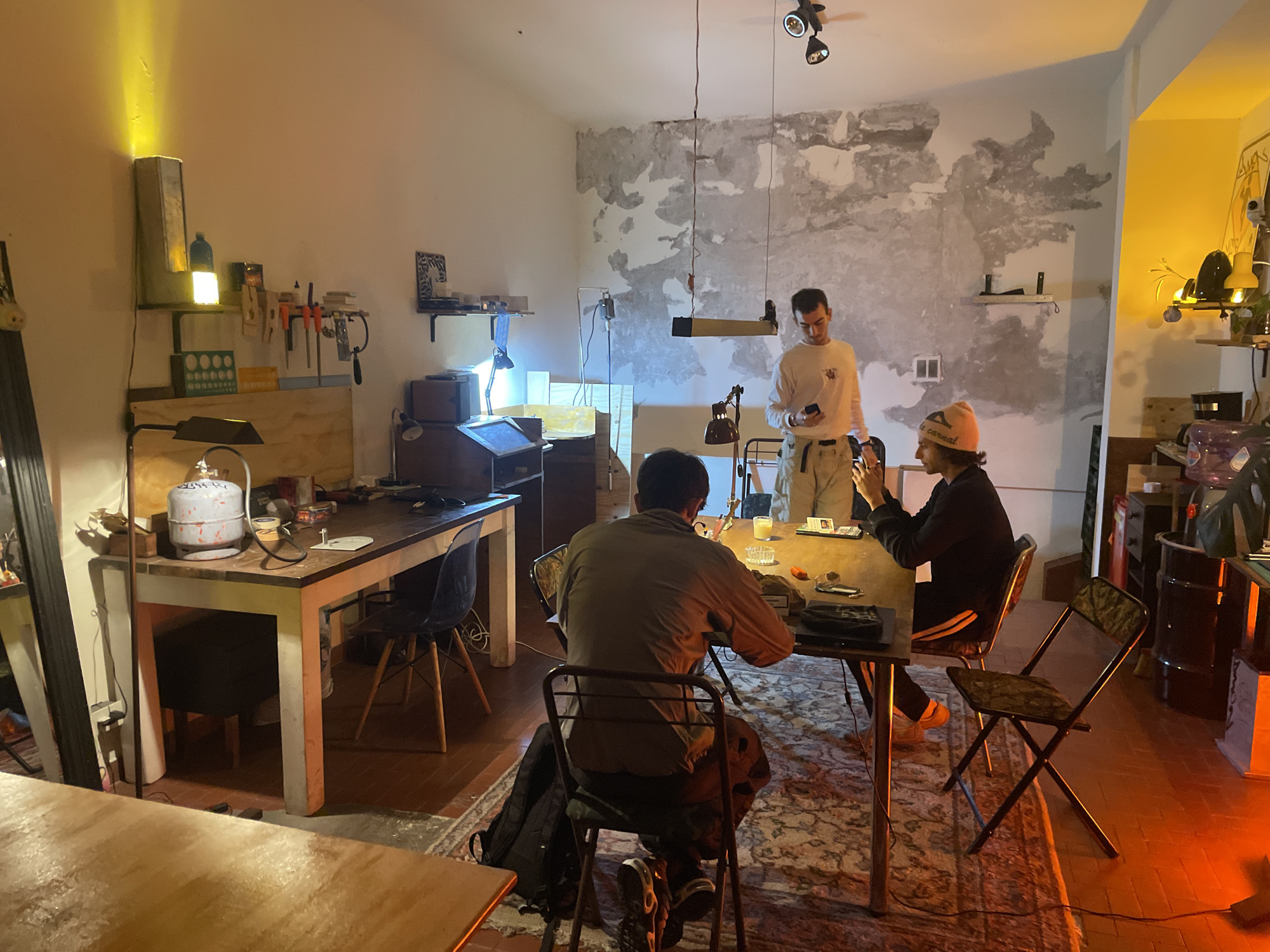 The image shows an interior space in the city of Buenos Aires, Argentina. Three individuals are present. One individual, standing, is using a mobile phone. Two seated individuals are at a table. The table has various objects on it including a candle and a container. The space has an industrial, workshop vibe, with worn walls, various tools, and different light sources. A rug covers part of the floor.