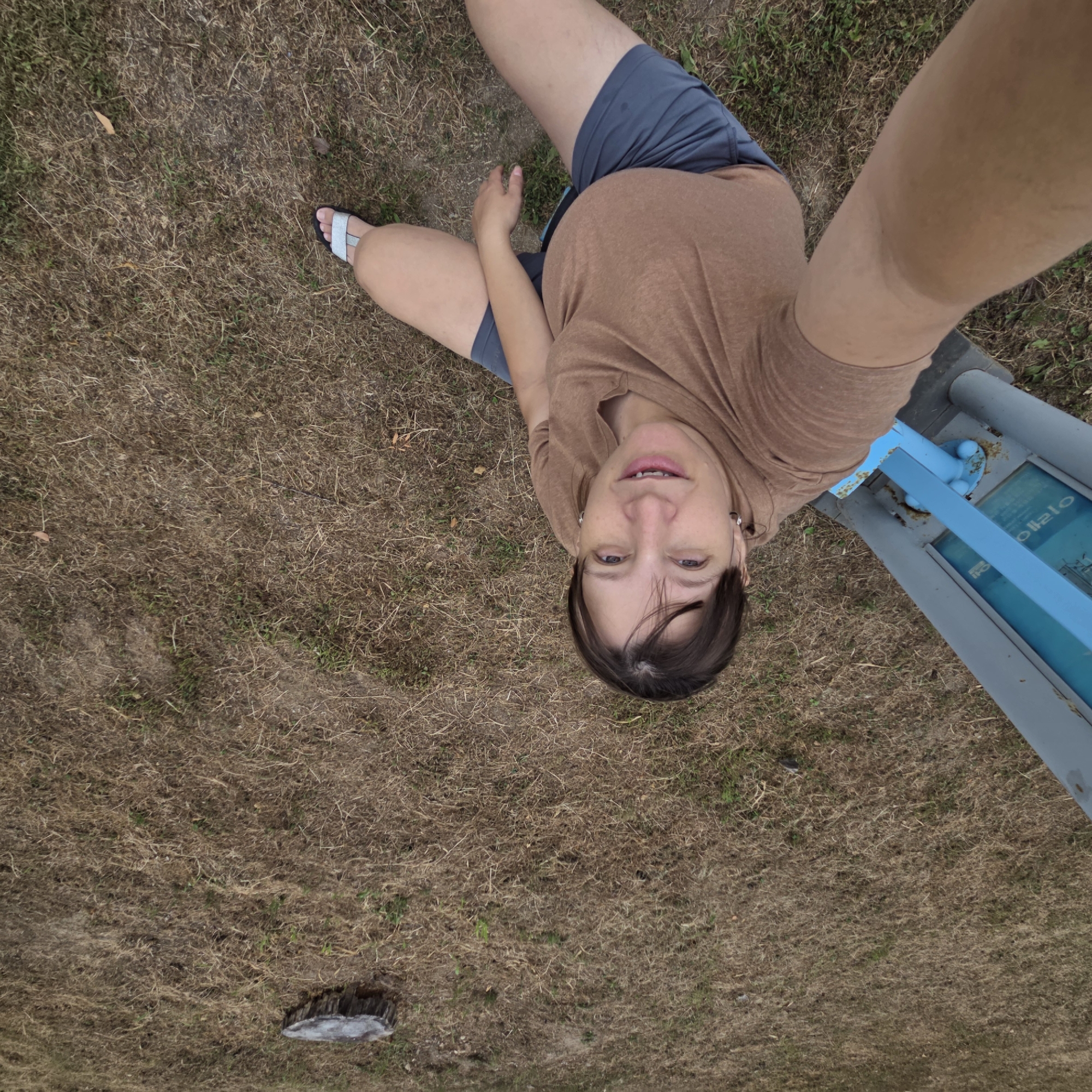 A woman is taking a selfie outdoors, near a blue metal pole structure that appears to be part of a park amenity. She is wearing a tan t-shirt and blue shorts, with white and silver sandals. The ground is covered with dry, light brown grass and some sparse green weeds. In the background, a tree stump is visible in the upper right portion of the frame. The image suggests a casual outdoor setting, possibly in a park or recreational area. The text on the blue structure is in Korean.