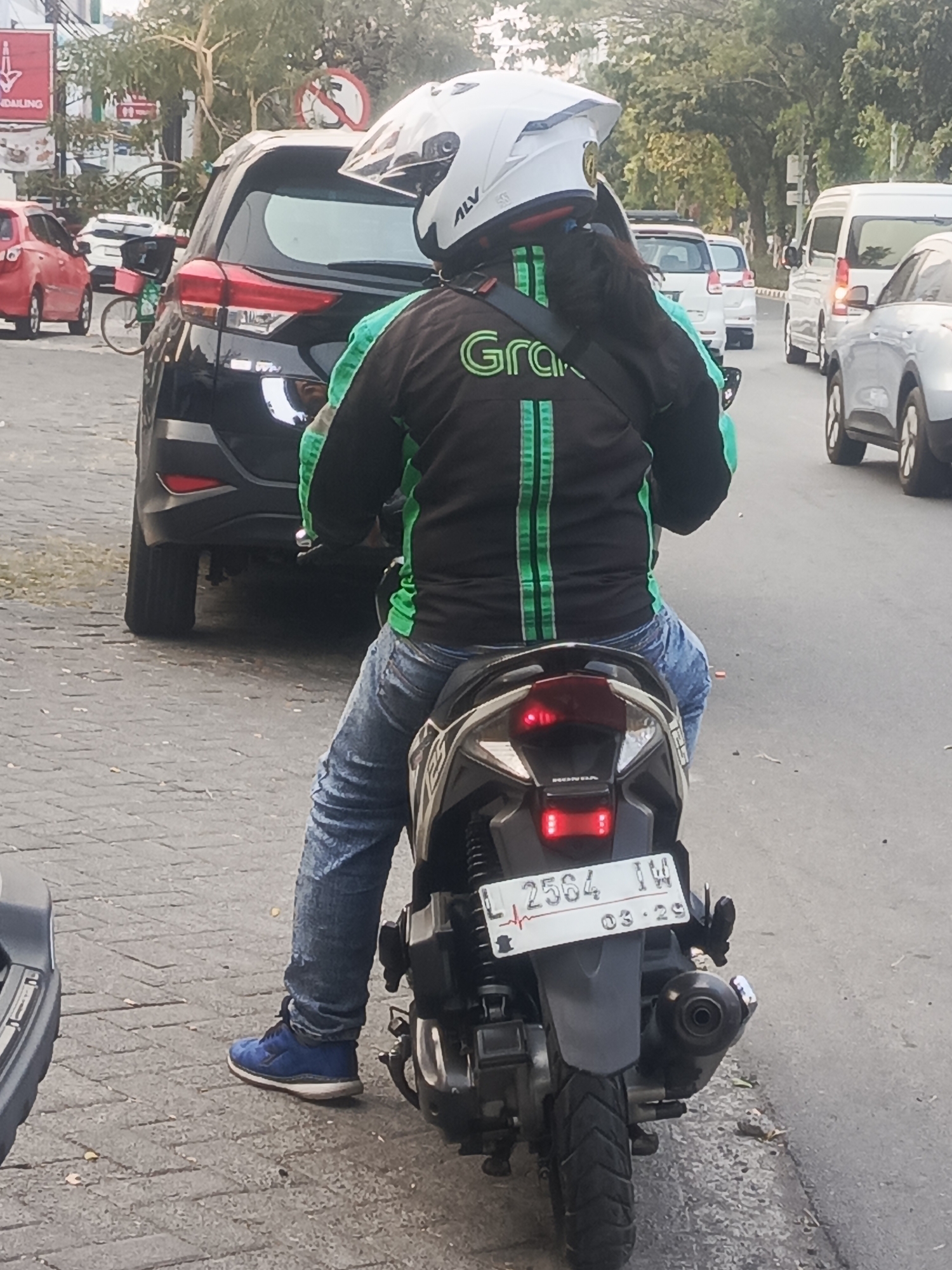 In Surabaya, Indonesia, a person wearing a white helmet and a black jacket with green trim is riding a motorcycle. The jacket has the "Grab" logo on it. The person is wearing blue jeans and blue shoes. The motorcycle's license plate reads "L 2564 IW" and has "03-29" printed on it. A black SUV is parked to the left of the motorcycle, and several other vehicles are visible in the background, including a white van and a red car. The scene appears to be on a street with a paved surface.