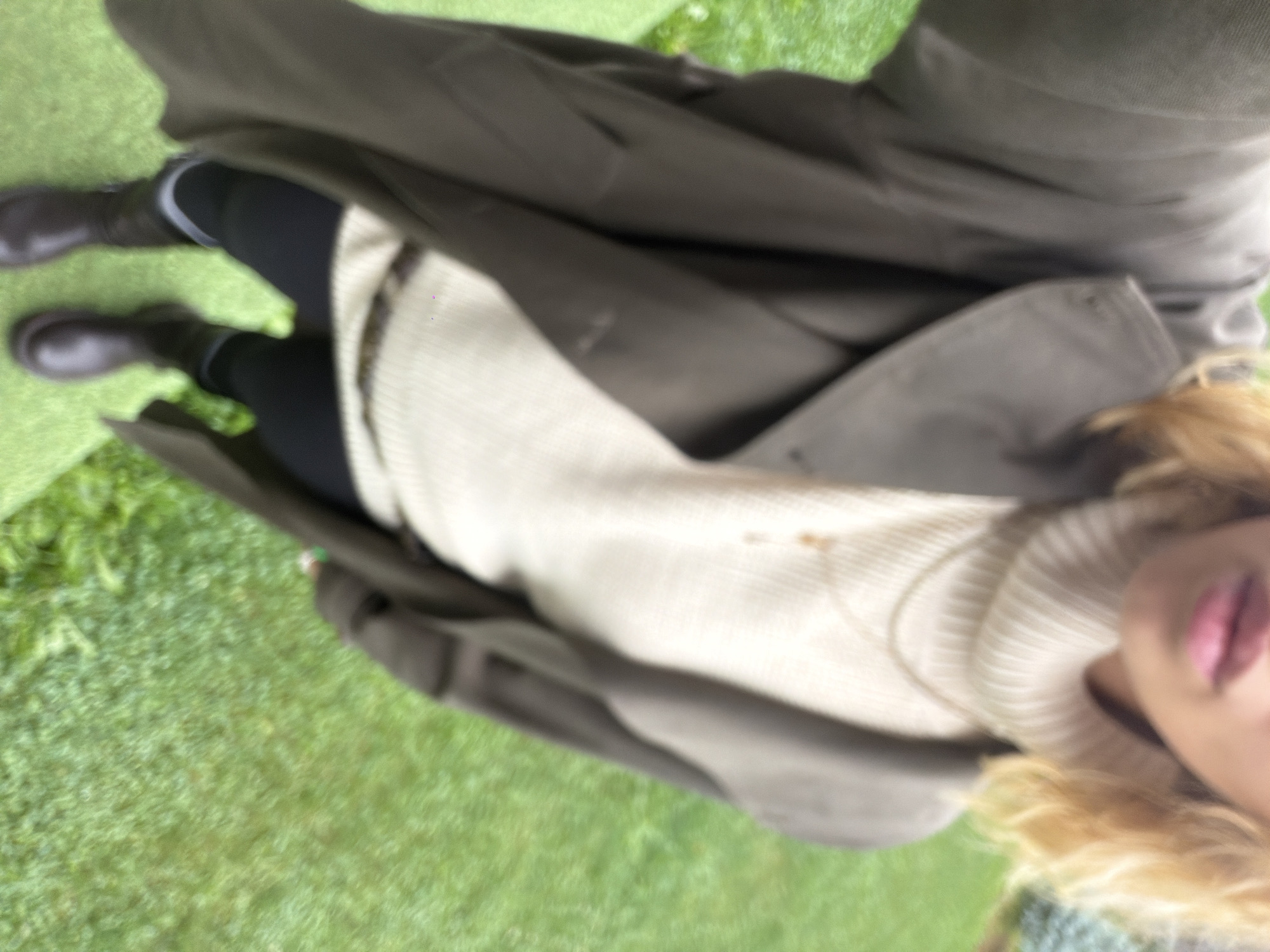 A person is standing on a green surface that appears to be grass or astroturf, possibly in an outdoor area like a park or yard. The person is wearing a dark olive green trench coat over a cream-colored turtleneck sweater. They are also wearing black leggings and dark brown boots. The background is slightly blurred, showing more green grass and some trees. The overall impression is a casual outdoor setting. The specific location in Kamenka, Russia, cannot be determined from the visual information provided.