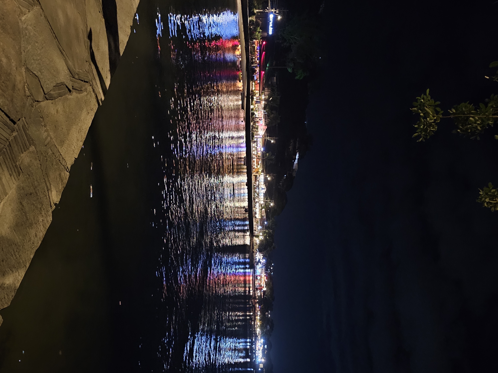 A long exposure photograph taken at night depicts a dark body of water reflecting a brightly lit area on the opposite shore. The water occupies the left side of the frame, and a concrete structure is visible at the top left corner. The reflected lights form vertical streaks and patterns of blue, red, and white. On the right side, the outline of trees is visible against the night sky. The scene is reminiscent of waterfront establishments and the reflection indicates the location is likely in the city of Armação dos Búzios, Brazil. The overall scene suggests a nighttime urban waterfront setting.