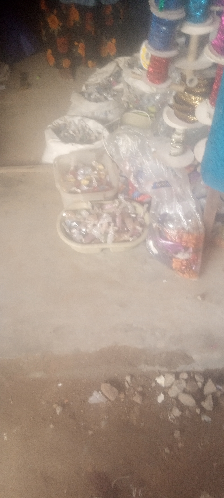 The scene depicts an outdoor market stall in Sabon Tasha, Nigeria. Visible are various bags and containers filled with wrapped candies, ribbons and other goods for sale. The scene is partially obscured by poor image quality and suggests a bustling marketplace environment.