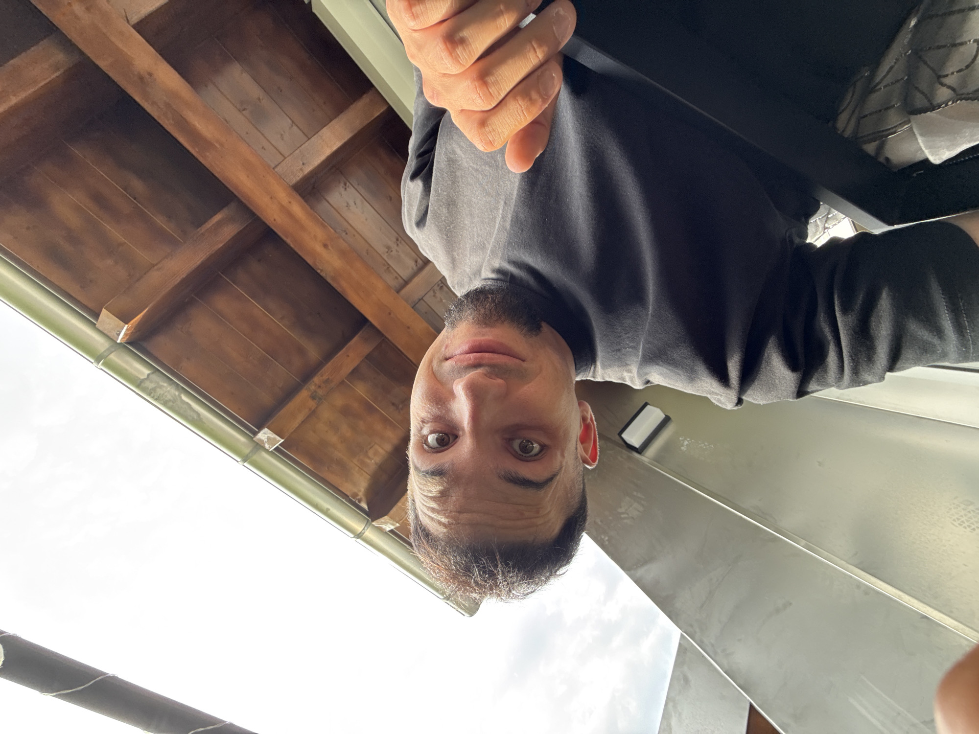 The image shows a low-angle, wide shot of a man with dark hair and a beard, looking directly at the camera with a surprised expression. He is wearing a dark t-shirt and his right hand is visible, extended towards the camera. Behind him, a wooden roof structure with exposed beams is visible, along with a silver-colored railing or architectural feature. The sky is overcast. There are no discernable landmarks that would place this image specifically in Garmisch-Partenkirchen, Germany.