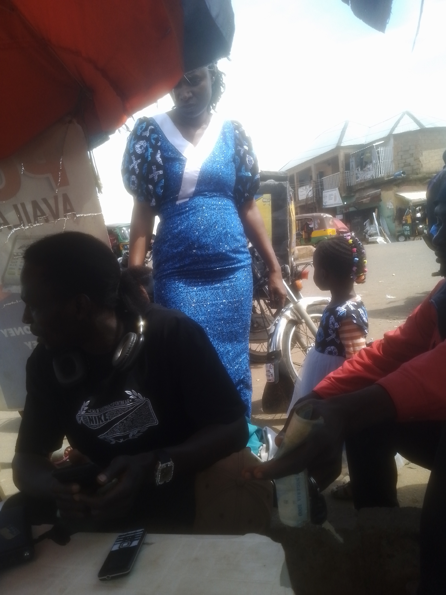 A group of people are gathered outdoors, possibly at a market or roadside stall, in Gwong, Nigeria. The scene appears to be during the day, with bright sunlight indicating fair weather.
In the foreground, a person wearing a black t-shirt with a Nike logo and headphones around their neck is seated, looking down at something in their hands. A mobile phone lies on the table in front of them. Beside them, another person in a red top is also seated, with their hands visible.
Behind them, a woman in a blue dress with a white neckline stands near a motorcycle. She appears to be looking in the distance. A young girl, wearing a dark dress with colorful beads in her hair, stands next to the motorcycle, looking to the side. The motorcycle has a few bags attached to it.
In the background, there are buildings that resemble shops or houses, suggesting an urban or semi-urban environment. A yellow and green auto-rickshaw is also visible, indicating local transportation. The overall impression is of a bustling, everyday scene in a Nigerian community. Some text is visible on the t-shirt of the person in the foreground, including "NIKE" and "CALIFORNIA SPORTSWEAR". Another piece of text, possibly currency, is being held by the person in the red top, showing "NIGERIA 1000".