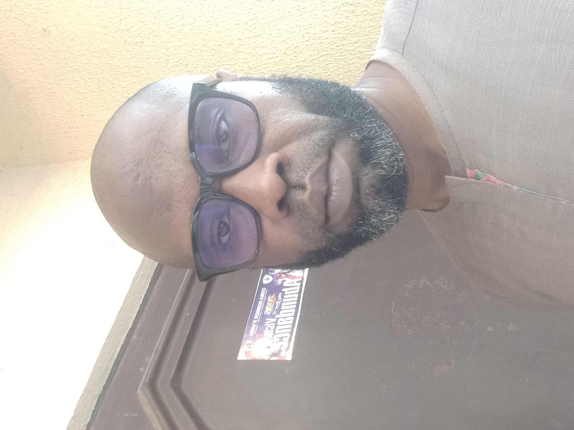 A close-up, slightly overexposed portrait features a man with dark skin, a bald head, and a dark beard flecked with grey. He wears black-framed eyeglasses with purple-tinted lenses and looks directly at the viewer with a neutral expression. He is dressed in a light-colored, possibly beige or light grey, textured shirt, with a hint of red and green patterned fabric visible at the neckline on the right side.
The background to the left and above his head consists of a textured, light yellow wall. To the right and below, a dark brown, paneled door is visible. A rectangular sticker is attached to the door, near the man's right cheek. Partially legible and reversed text on the sticker includes "MAY 5 & SUNDAY", "SOUNDBUNCH", and "AFRI". The image is brightly lit, likely by natural light, which causes some glare on his forehead and glasses. The setting is an everyday environment in Barden Yana, Nigeria.
