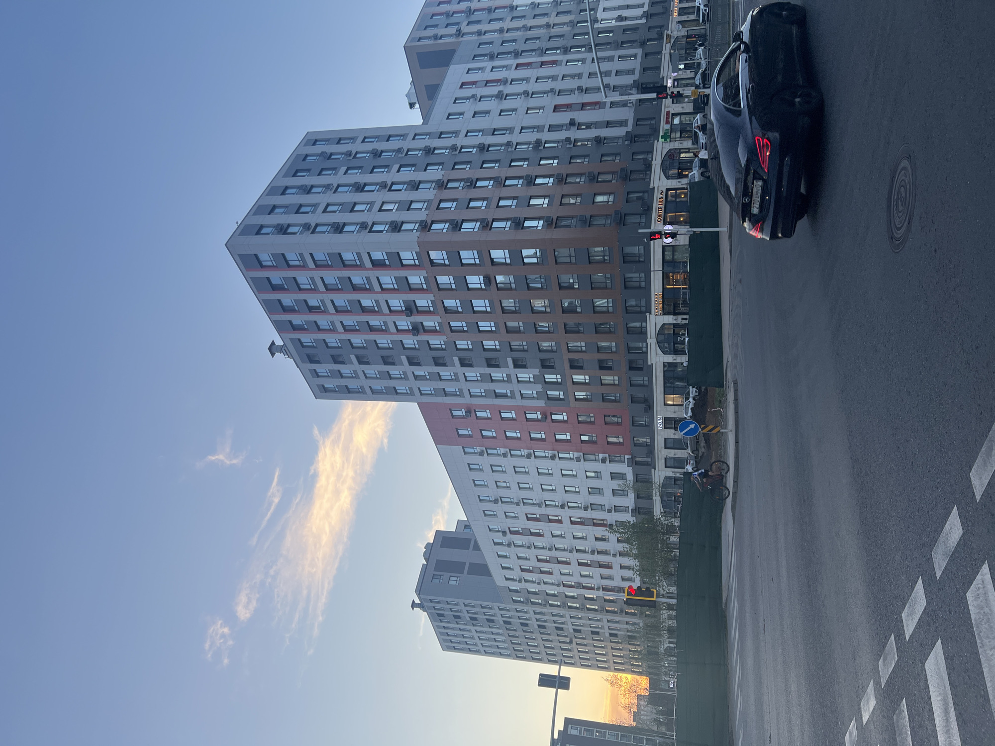 A street scene in Astana, Kazakhstan features a modern apartment building with a segmented facade of grey, white, and red panels. The building has numerous windows and external air conditioning units. In the foreground, a black car, likely a Mercedes-Benz, drives through an intersection with a visible license plate. A cyclist is also present, riding on the sidewalk. Traffic lights are visible, with one showing a red light. The sky is a clear blue with wispy clouds illuminated by the setting sun. The street is paved and marked with pedestrian crossings. Green fencing is present along the side of the road.