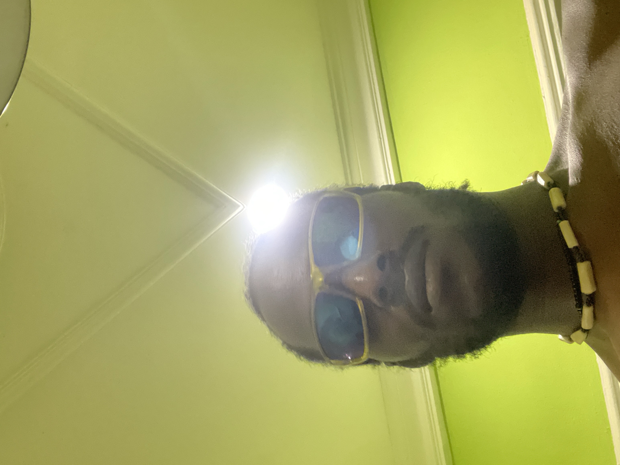 A medium shot depicts a man with dark skin, a beard, and sunglasses. The lighting in the photograph is bright, with a glare reflecting off the glasses. The background consists of a light yellow wall, a white door, and a strip of green wall. The man is wearing a beaded necklace. The location is unclear, but is said to be in the city of Abuja, Nigeria. The photograph is taken from a low angle, looking upwards at the man.