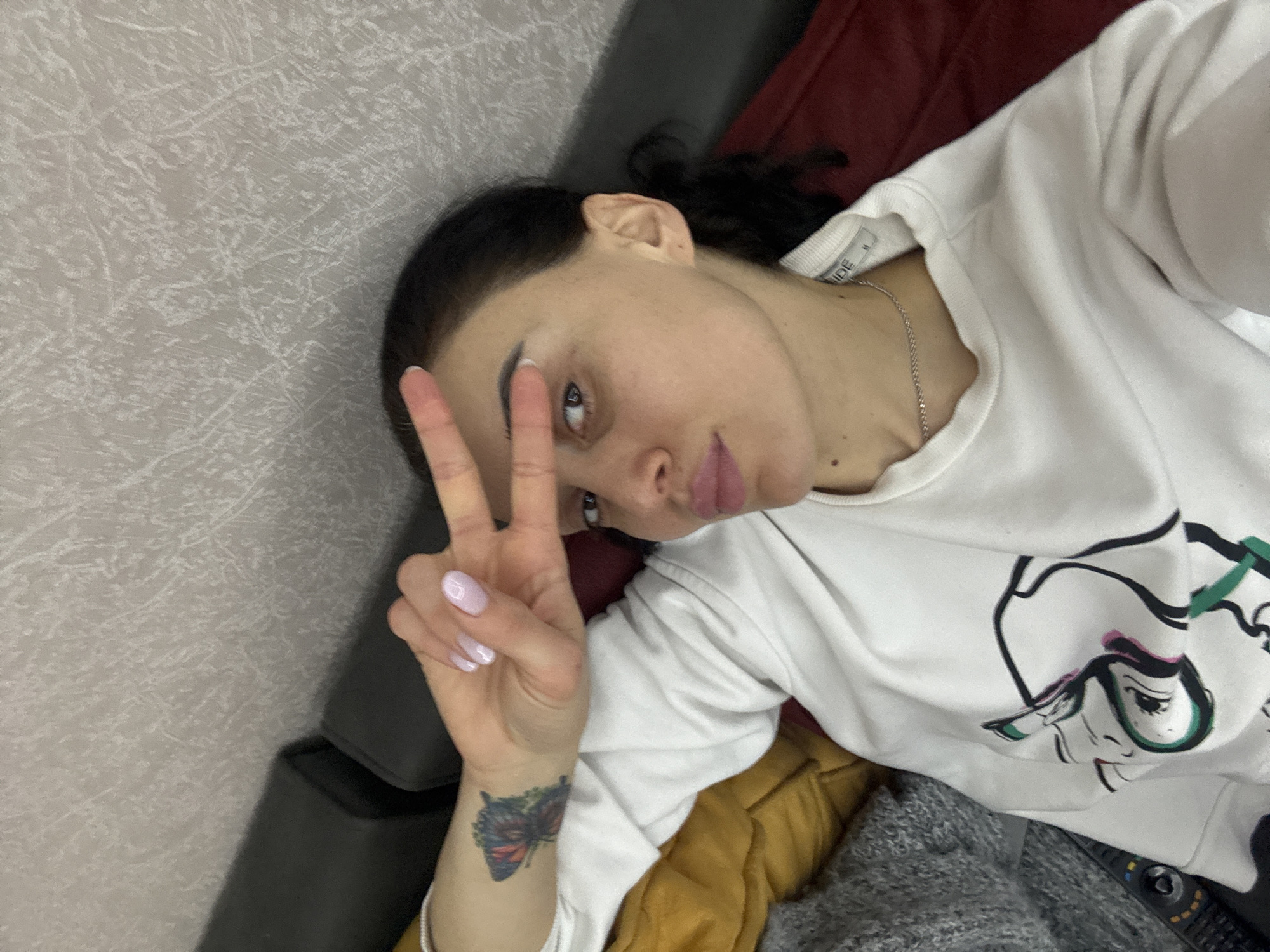 A woman is lying down indoors, oriented diagonally in the frame. She is making a peace sign with her left hand. The woman has dark hair pulled back and light skin. She is wearing a white sweatshirt with a cartoon graphic and a silver chain necklace. A butterfly tattoo is visible on her left wrist. The background is a textured grey wall and the edge of what appears to be a dark-colored couch or cushion. The setting is likely indoors, but the location within Tbilisi, Georgia cannot be determined from the visual information provided. The picture appears to be a selfie.
