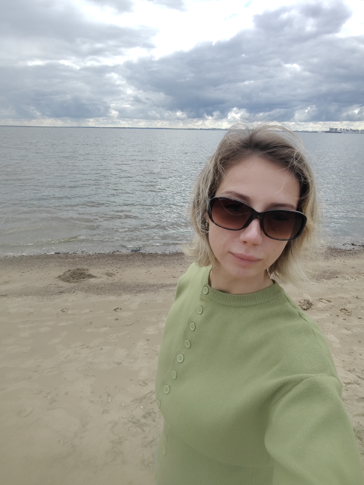 A woman with short blonde hair and sunglasses is standing on a sandy beach, facing the camera. She is wearing a light green long-sleeved shirt with buttons. Behind her is a body of water, likely the Gulf of Finland, with a cloudy sky overhead. In the distance, on the horizon, there are some buildings and structures visible, suggesting an urban area. The scene appears to be taken in the city of Saint Petersburg, Russia, given the characteristic landscape of the Gulf of Finland and its proximity to the city's infrastructure. The overall mood is neutral, with the focus on the subject and the vastness of the natural environment.