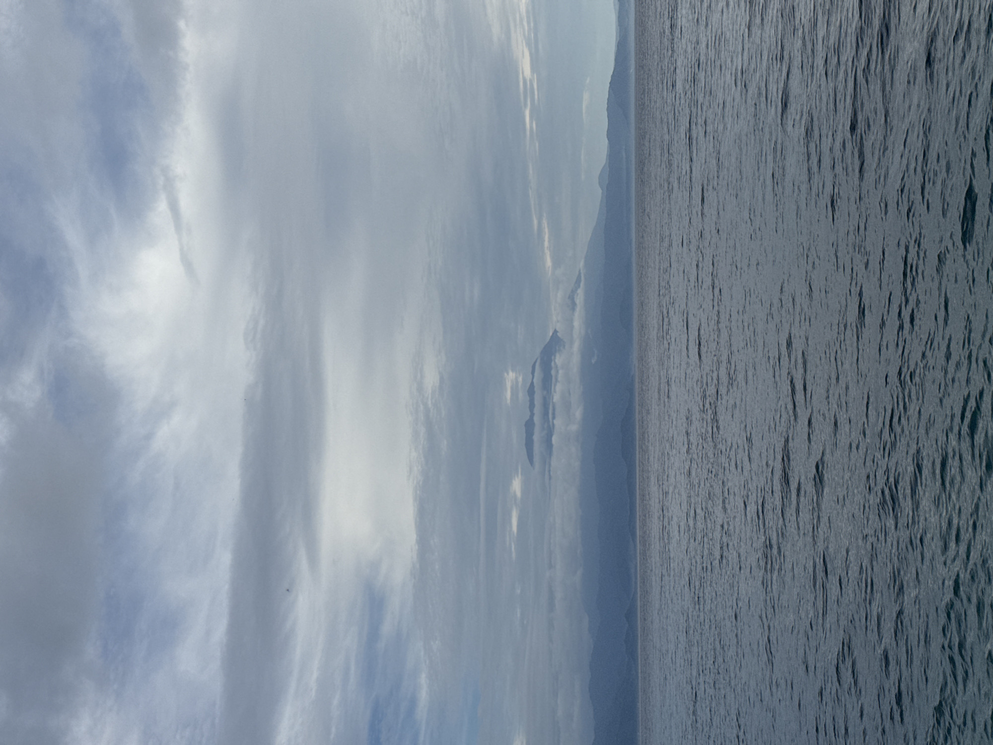 The image shows an outdoor scene with a body of water on the right side and a cloudy sky on the left. In the background, there are mountains partially obscured by clouds. The water appears choppy, suggesting a breeze. No people or specific objects are visible.