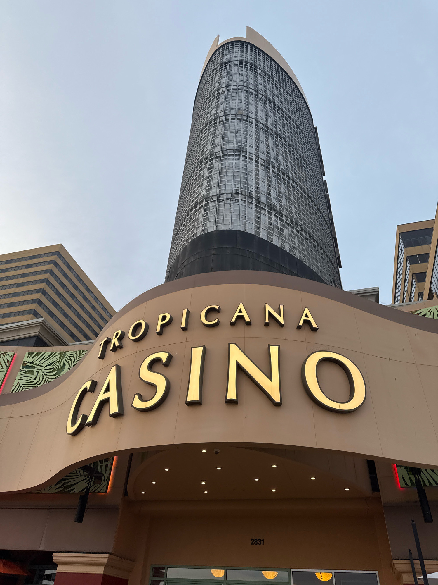 The image shows the Tropicana Casino and Resort in Atlantic City, New Jersey, United States of America. The photo is taken from a low angle, looking up at the hotel's facade. The curved entrance to the casino is prominent, with large, gold-colored letters spelling out "TROPICANA CASINO." Above the casino entrance, a modern, cylindrical skyscraper with a glass facade towers into the sky. To the left and right of the main tower are other buildings, also with glass windows. The sky is overcast and pale. The entrance area below the sign is covered by a canopy, illuminated by recessed lighting. A small sign with the numbers "2831" is visible near the bottom of the entrance area. The overall scene depicts a large, contemporary entertainment complex.