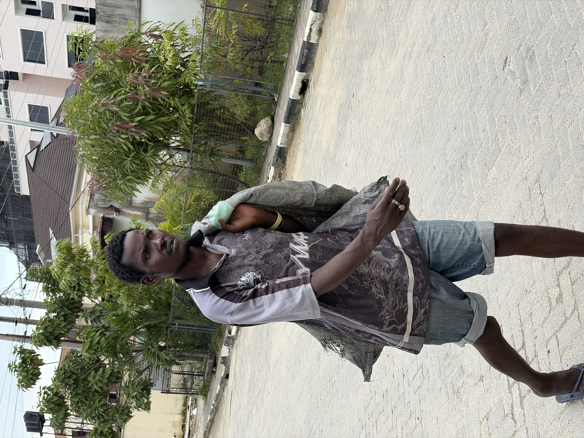 A full shot shows a Black man walking on a paved street in Igboefon, Nigeria, carrying a grey and green bag over his left shoulder. He is wearing a short-sleeved grey and black polo shirt with a graphic pattern and light blue denim shorts. The man is walking from left to right, with his head turned towards the right side of the frame, looking into the distance. He is wearing dark blue flip-flops. In the background, there are trees, a fence, and buildings. The street has a paved surface with a black and white painted curb. The sky is overcast.