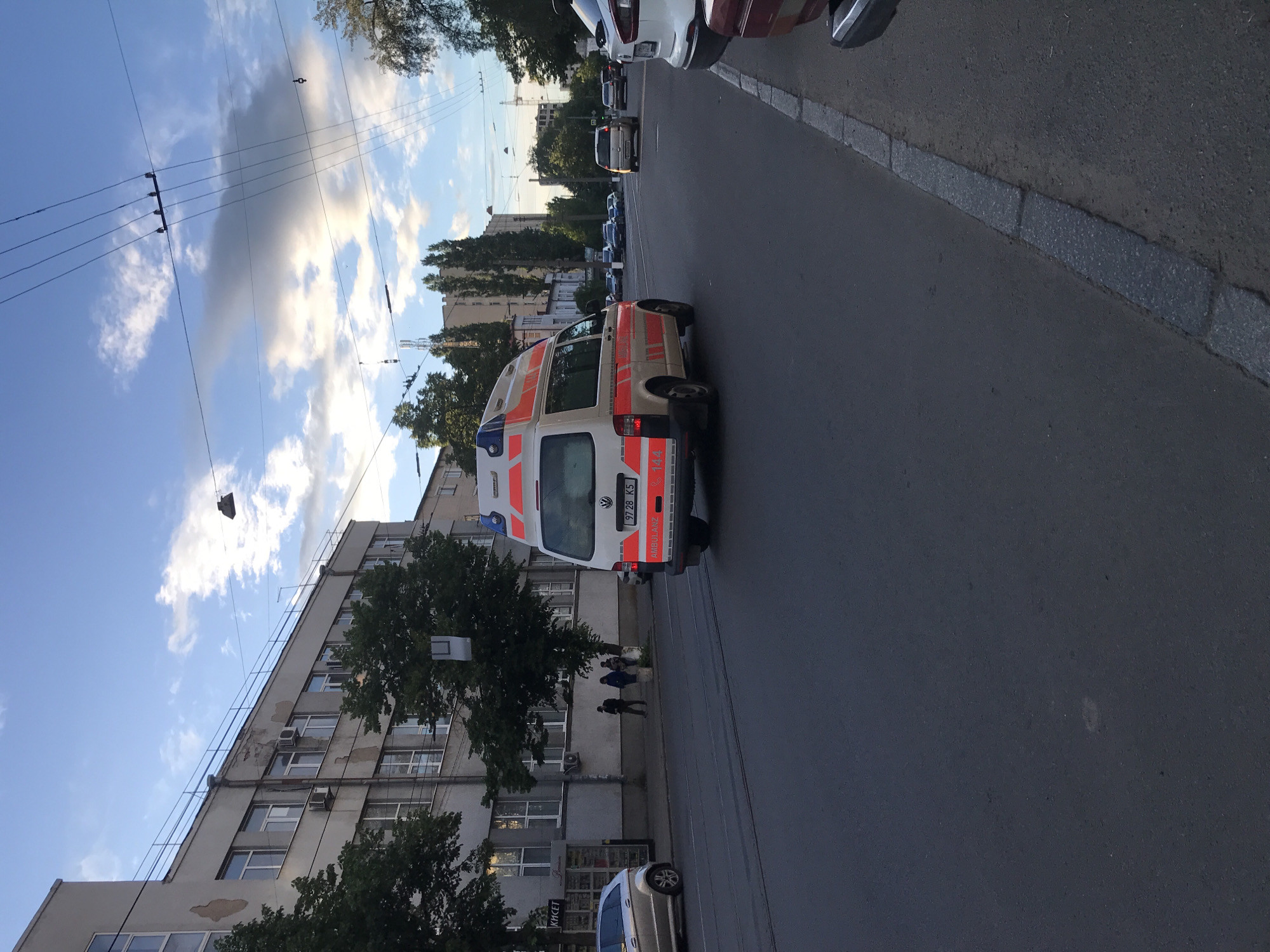 An ambulance vehicle, white with orange striping, is driving on an asphalt city street in Kharkiv, Ukraine. The license plate on the ambulance reads "9728 K5." The street is lined with parked vehicles. The building on the left has multiple windows and air conditioning units. There are people visible near the building. Overhead, there are power lines. The sky is partly cloudy.