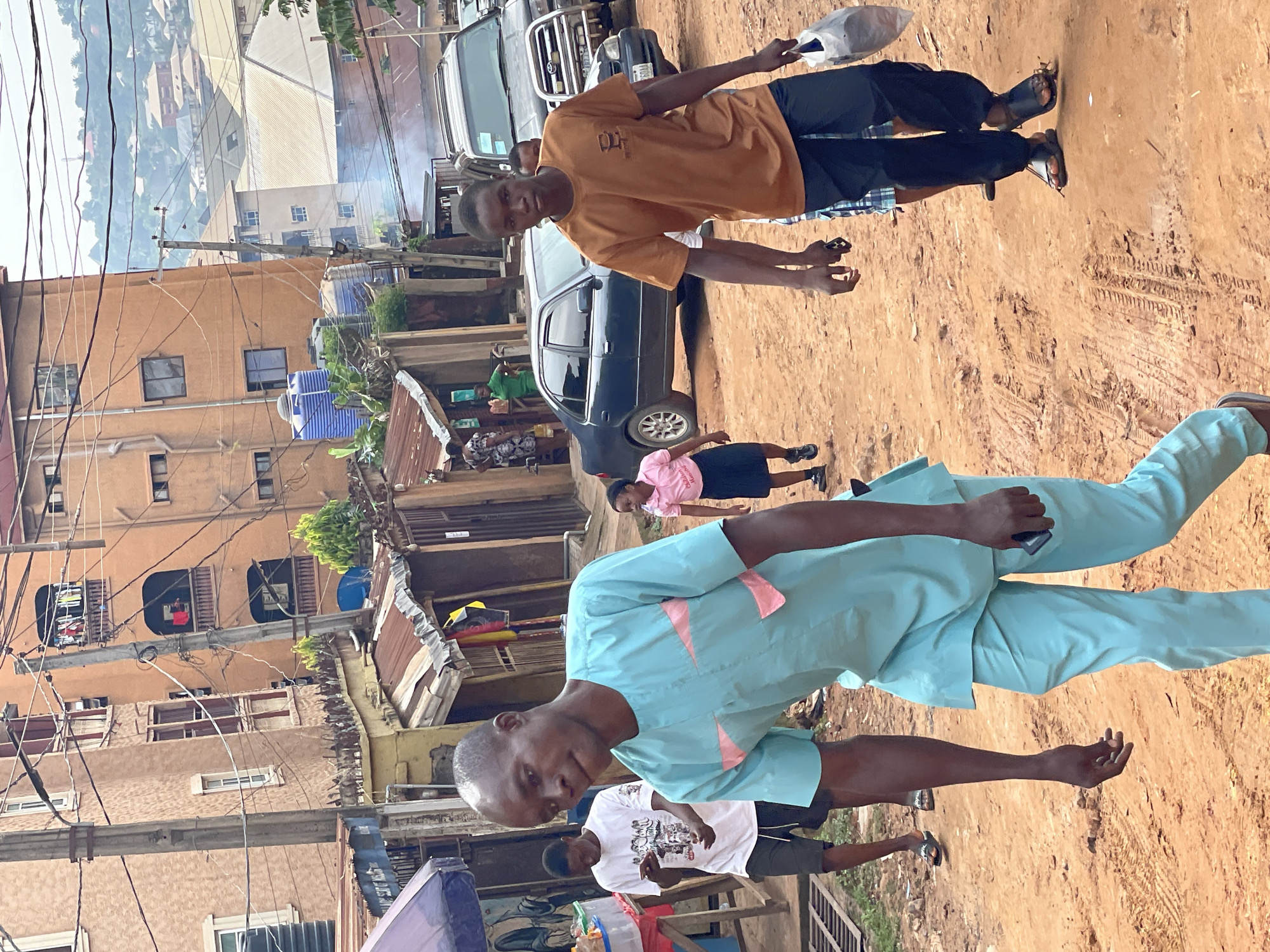On a dirt road in Onitsha, Nigeria, a man wearing a light blue outfit with pink accents walks towards the camera while holding a cell phone. Behind him is a man wearing a white t-shirt. In the background, a young girl in a pink shirt walks behind a dark blue sedan. A tall man in an orange shirt stands behind a silver SUV. The background includes closely placed buildings with weathered facades, exposed wiring, and a view of a cityscape in the distance.