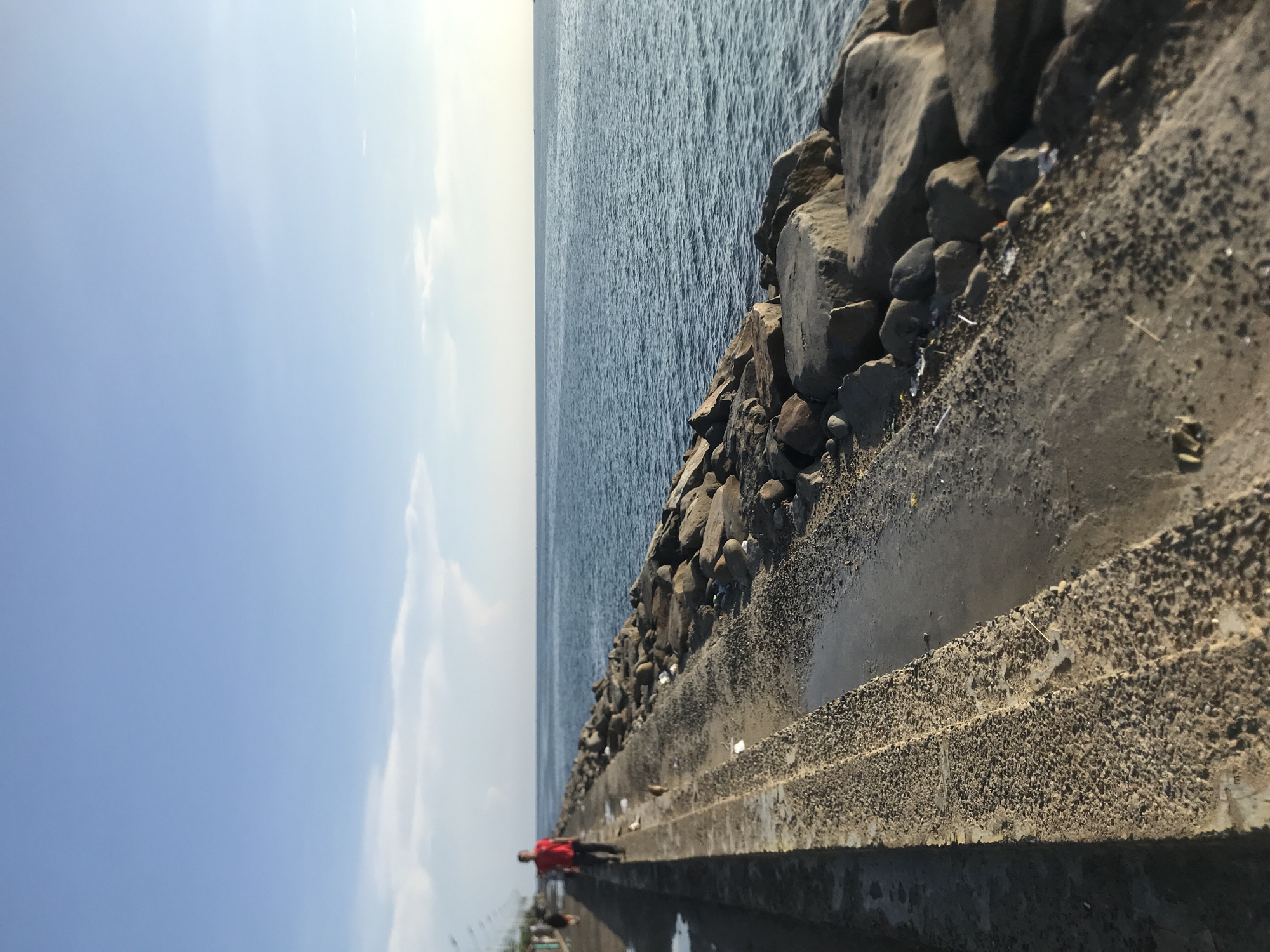 The image shows a view from a pier in Lhokseumawe, Indonesia. The pier is made of concrete and lined with rocks to protect it from the sea. The ocean is visible on the right, and the sky is on the left. There is a person wearing a red shirt lying on the pier. The pier is covered in debris and water.