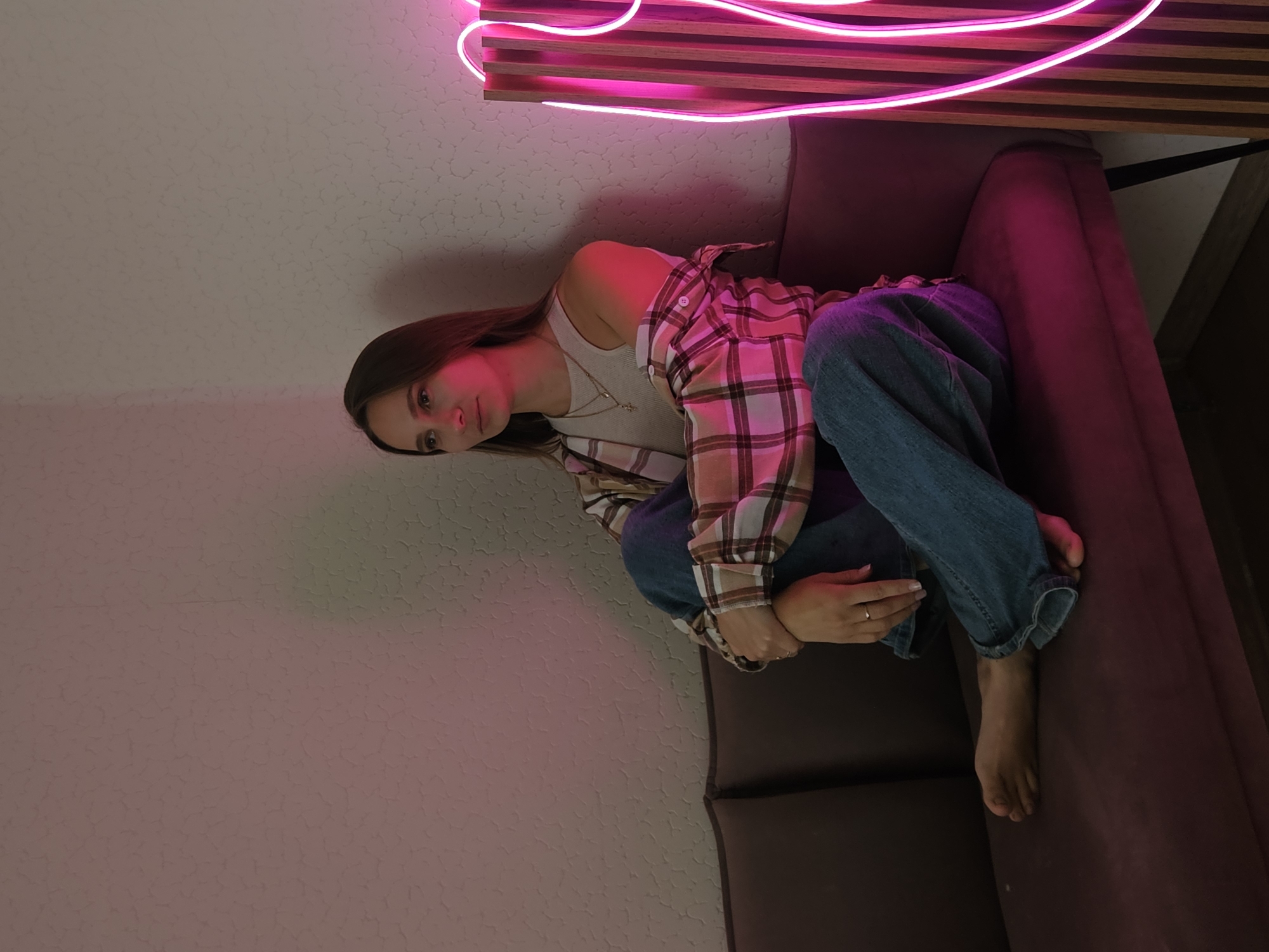 The image depicts a young adult female, approximately 20-30 years of age, seated on a dark brown upholstered sofa in an indoor setting in Chaikovsky, Russia. She is positioned with her knees drawn up and arms crossed over them, looking towards the left side of the frame with a neutral expression. Her long, straight brown hair is down. She wears a light-colored tank top, an open plaid button-up shirt in brown, beige, and pink tones, and blue denim jeans; her bare right foot rests on the sofa. Two layered necklaces and a ring on her right ring finger are visible. The background features a light-colored, textured wall. A prominent pink neon light fixture, partially visible on the right, casts a strong magenta glow across the scene, impacting the woman, the sofa, and the wall. A subtle green light is also evident on the upper right portion of the wall.
