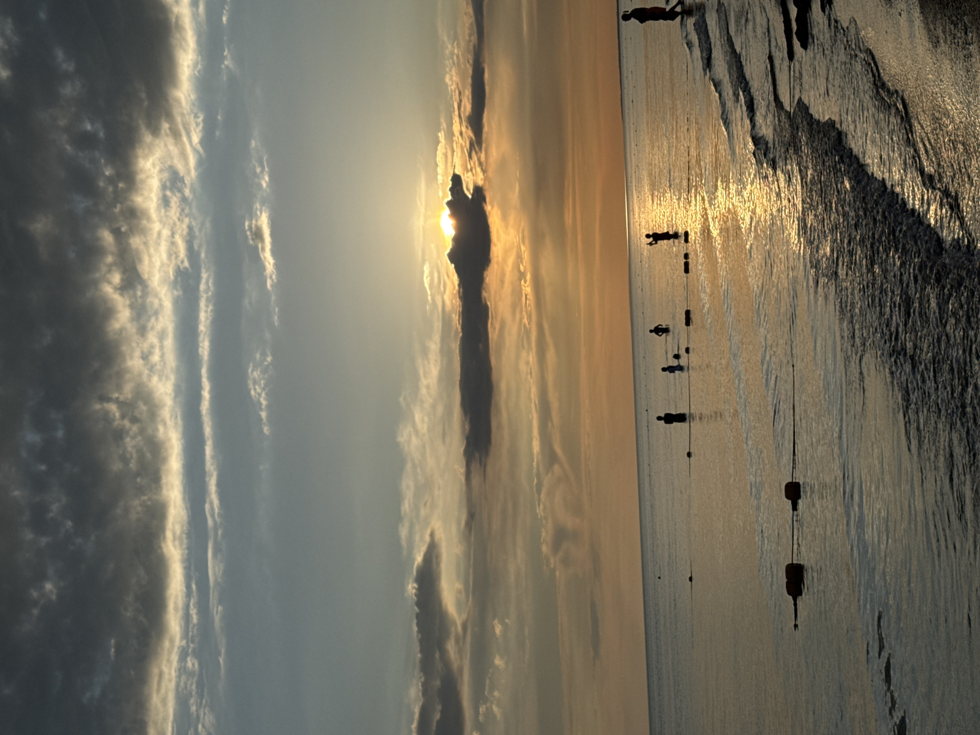 The image depicts a sunset scene at a beach in Vityazevo, Russia. Silhouetted figures of people are visible in the shallow water and on the shore. The sun is low in the sky, partially obscured by clouds, casting a warm golden glow on the water and sky. The sea is calm with gentle waves lapping at the sandy shore. Buoys are visible in the water, marking a swimming area. The sky displays a gradient of colors from deep grey clouds at the top to warm oranges and yellows near the horizon.