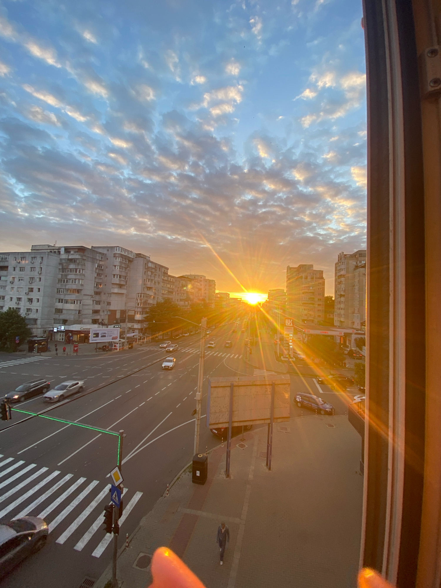 The image captures a sunset in Iasi, Romania, viewed from a window frame on the right. The sky is filled with a dramatic display of clouds, illuminated by the setting sun, which casts long rays across the scene. Below the sky, a city street is visible, lined with apartment buildings that appear to be from the Soviet era, characterized by their concrete facades and repetitive window patterns. Traffic can be seen on the multi-lane street, including cars and what appear to be buses. Pedestrians are present on the sidewalks and at a crosswalk. A gas station is also visible on the right side of the street. The overall impression is of an urban environment at dusk, bathed in the warm glow of the setting sun.