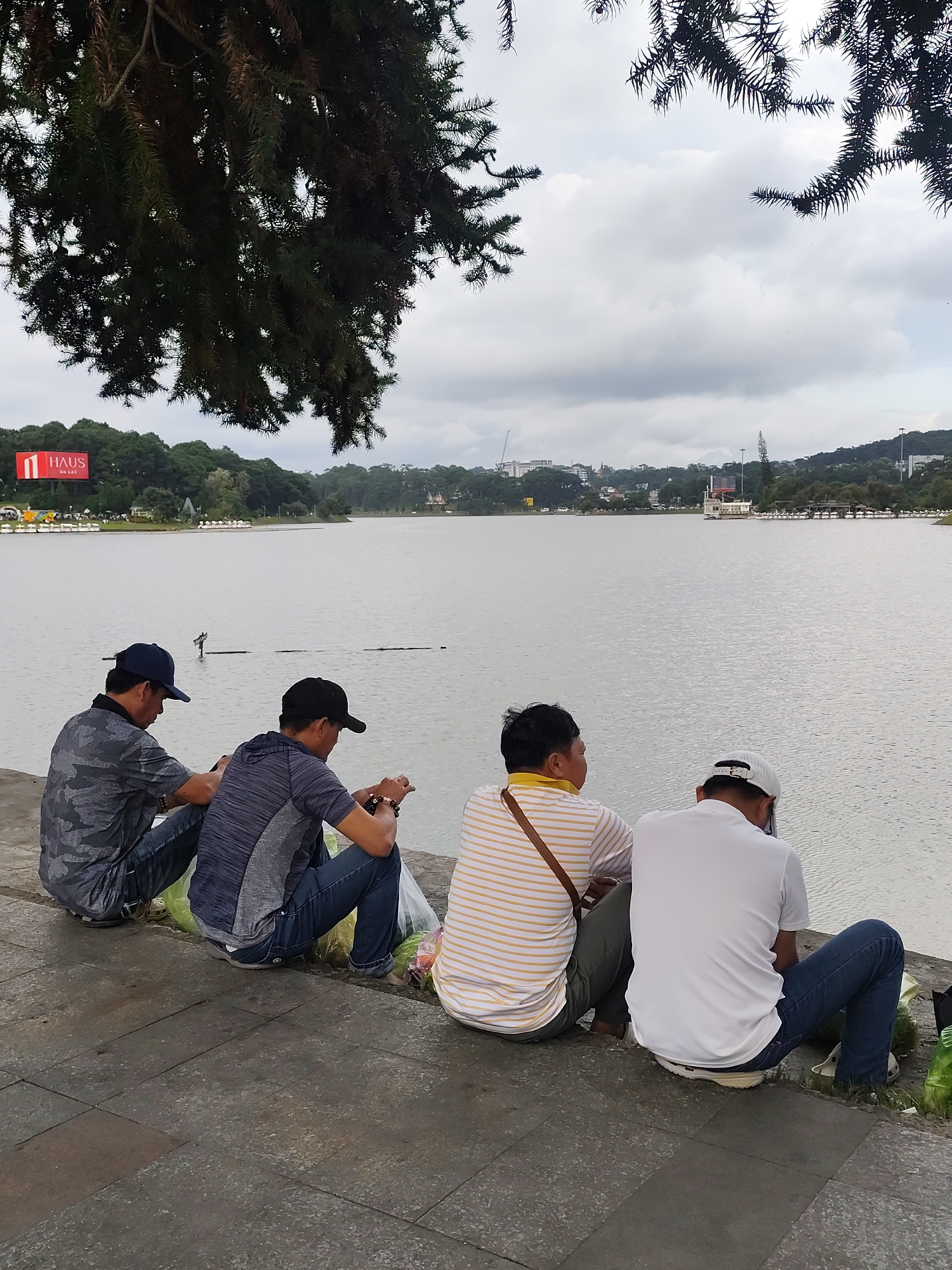 Four individuals are seated on a paved walkway adjacent to a large body of water. Two men in the foreground are wearing blue jeans and baseball caps, looking down. The man on the left is wearing a camouflage patterned shirt, and the man to his right is wearing a dark grey striped shirt. The third individual from the left is wearing a yellow and white striped shirt and is facing away from the camera. The fourth individual, on the far right, is wearing a white polo shirt and blue jeans, with a white baseball cap, also facing away from the camera. They are all seated with their legs extended. Several plastic bags, some appearing to contain produce, are on the ground near the individuals. In the background, across the water, is a city skyline with trees, buildings, and a large sign with the word "HAUS" on it. The sky is overcast.