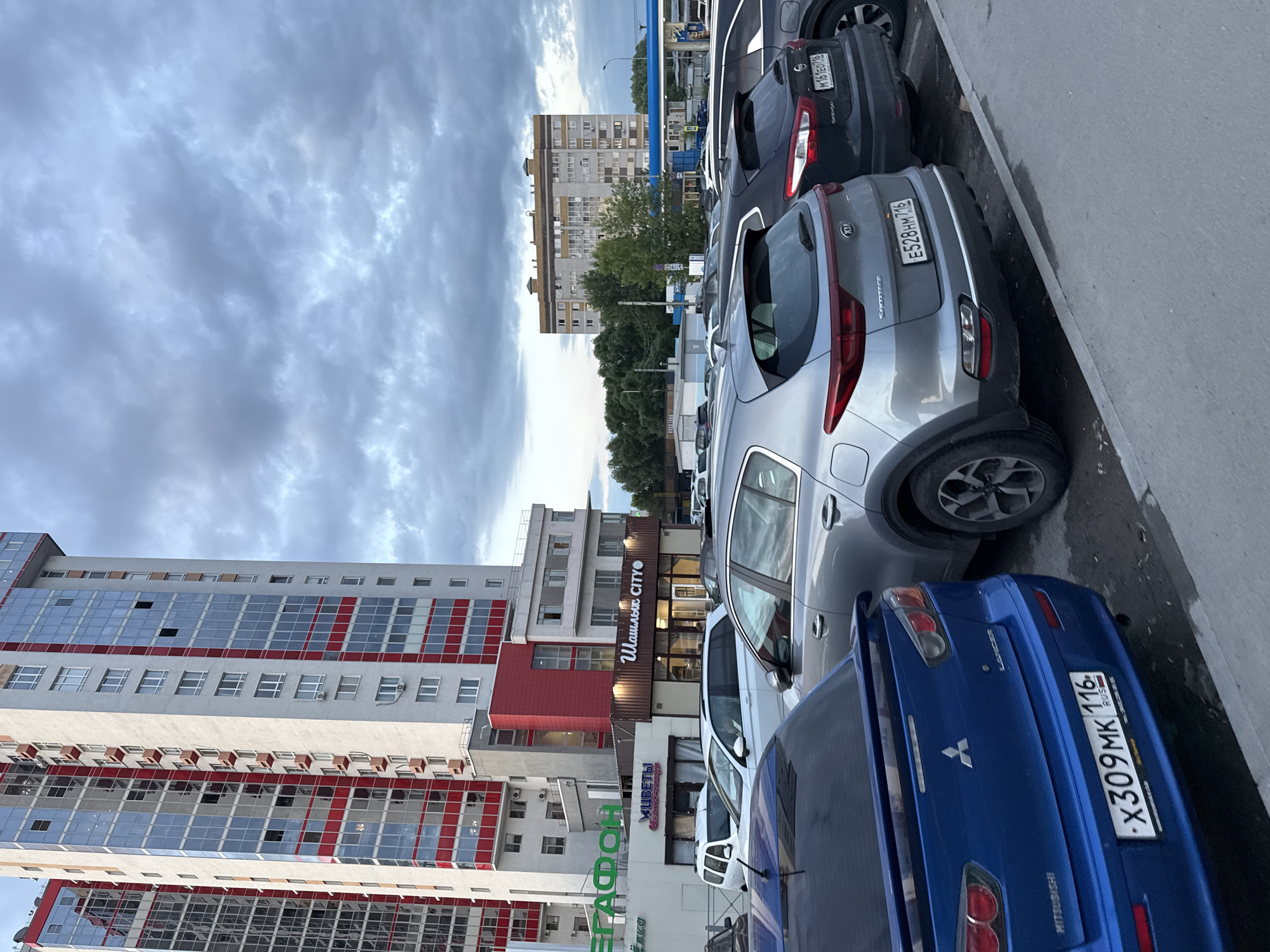An outdoor, eye-level, full shot captures a street scene in Kazan, Russia, under an overcast sky. In the foreground, several cars are parked, including a blue Mitsubishi Lancer with the license plate "X309MK 116 RUS," a silver Kia Sportage with the license plate "E528HM 716," and a dark-colored Nissan Qashqai with the license plate "M161EO 716." Further back, a white SUV is also visible.
Dominating the left side of the frame is a tall, modern apartment building with a white facade accented by red vertical stripes and numerous windows. To the right of this building, a row of shops with signage is visible, including "ЕРАФОН," "ЦВЕТЫ" (Flowers), and "Шашлык CITY" (Shashlik City).
In the background, another, older apartment building with a beige facade and multiple windows stands amidst a backdrop of green trees. A blue awning of a gas station can also be seen in the distance. The overall impression is a typical urban setting, likely a residential or commercial area within Kazan. No people are discernible in the image.