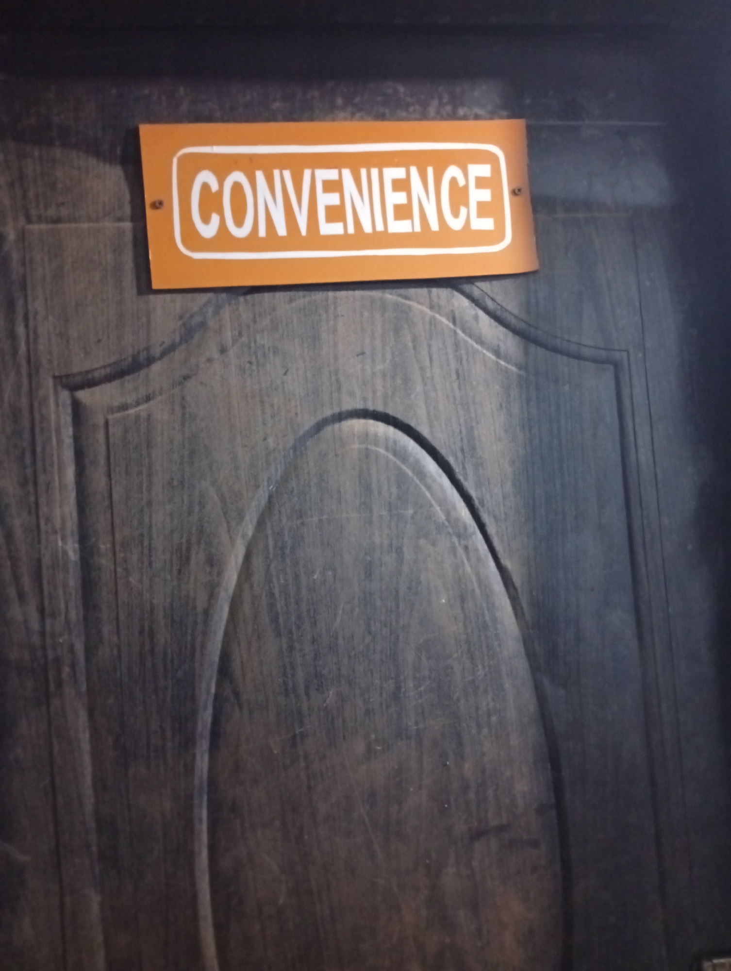 A dark, wooden door features a recessed, rounded panel design. Affixed to the door's upper portion is a rectangular orange sign with the word "CONVENIENCE" displayed in white block letters. The sign is mounted to the door with what appear to be metal screws. The location is presumed to be Adankolo, Nigeria.