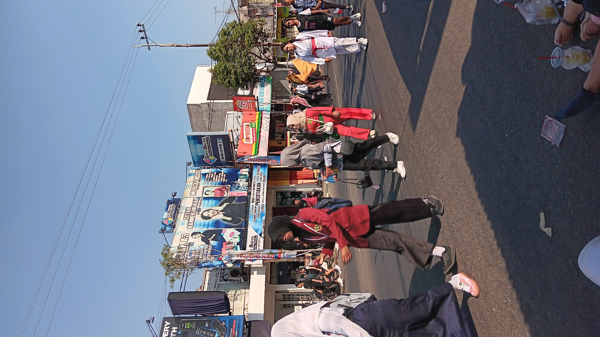 The image captures a street scene in Tanjung Anom, Indonesia, likely during a parade or procession. In the foreground, several individuals are walking along the asphalt road. On the left, a person wearing a dark blue skirt and a white headscarf is partially visible, walking away from the camera. Next to them, a person in a maroon blazer and dark pants, wearing a black hijab, is the central figure in the foreground, looking to their right and holding something in their left hand. To their right, a person in a grey hijab and black pants is walking, holding a paper cup with a straw in their left hand. Further to the right, a person in a red outfit and a light-colored hijab is also walking. In the middle ground, a group of people are walking, including a person in a white karate-like uniform with a red belt, and others in various attire. Behind them, a large banner advertising "DANI DIGITAL" and various printing services is prominently displayed on a building. Other signs and structures line the street, suggesting a commercial area. The sky is clear and blue, indicating daytime. The ground is paved with asphalt, and shadows are cast by the people and structures, suggesting the sun is positioned to the right of the frame. Several plastic cups with straws are visible on the ground in the lower right corner, along with what appears to be discarded packaging. The overall atmosphere suggests a public event taking place on a sunny day.