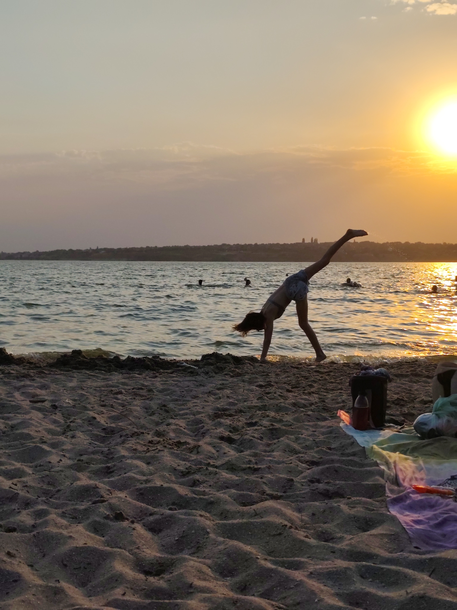The image depicts a sunset over a body of water, likely a river or a sea, adjacent to a sandy beach. In the foreground, the textured sand is visible, with some ripples and indentations. Further back, several people are in the water, appearing to swim or relax.
The main subject of the image is a young person in silhouette, performing a cartwheel on the wet sand at the water's edge. Their body is extended, with one leg high in the air, creating a dynamic pose against the backdrop of the setting sun. The sun itself is a bright, warm orb positioned in the upper right quadrant of the image, casting a golden glow on the water's surface and creating reflections.
On the beach to the right of the cartwheeling figure, there are some beach accessories, including a dark bag, a bottle, and a colorful blanket spread out on the sand. In the distance, a faint outline of land with trees and structures can be seen along the horizon. The sky is a gradient of soft colors, transitioning from a pale yellow near the sun to pinks and purples further away, with a few wispy clouds.
The scene conveys a sense of leisure and activity on a beach during a beautiful sunset. The location is described as Mykolaiv, Ukraine, suggesting this is a waterfront area within or near the city.