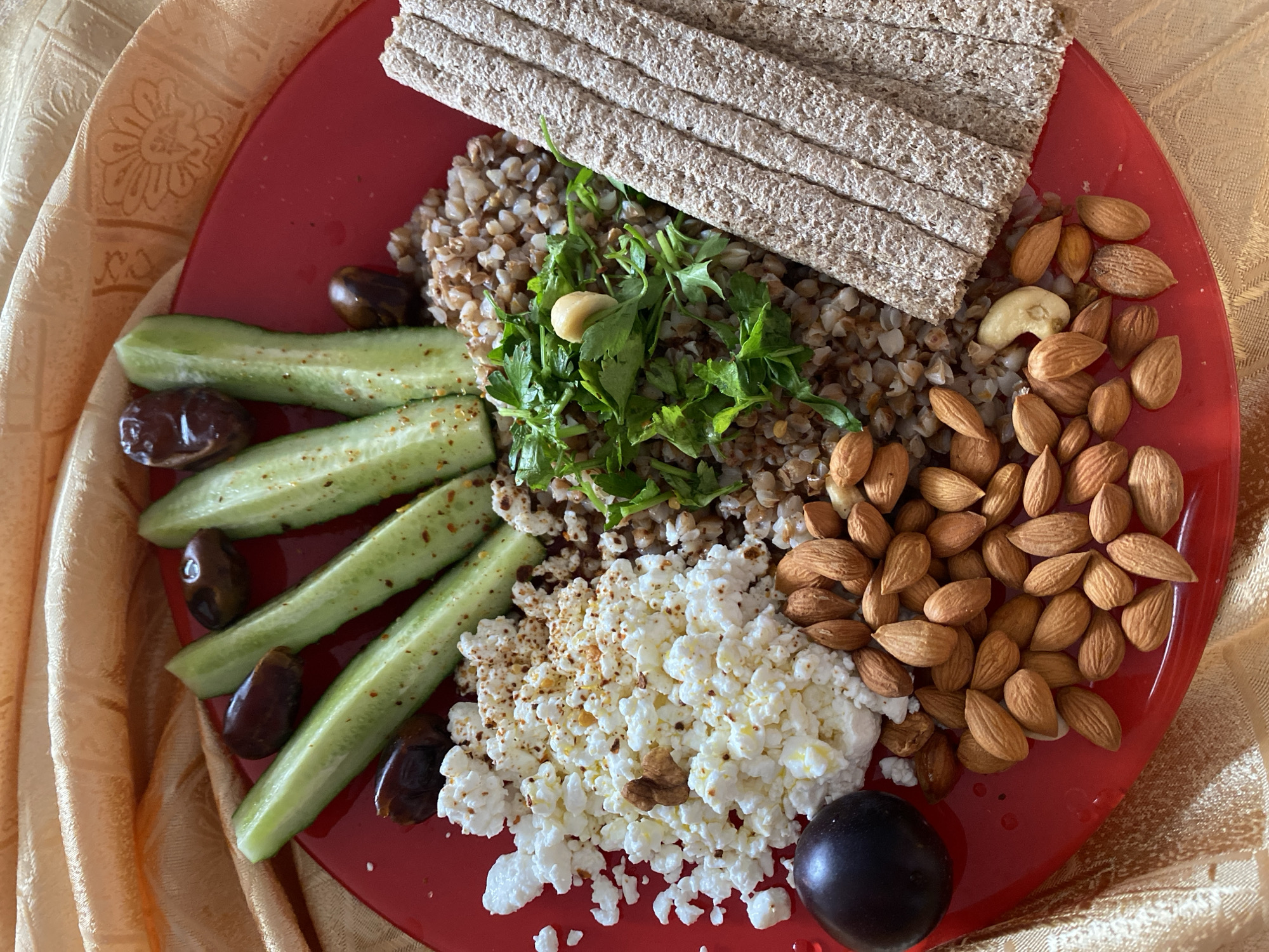 A close-up, top-down shot shows a red plate filled with food on a textured, gold fabric surface. The plate contains several items: sliced cucumbers seasoned with red pepper flakes, dark brown dates, a pile of white cottage cheese sprinkled with spices, a portion of cooked buckwheat, chopped fresh parsley, almonds, a single dark plum, and several flatbread crackers. The gold fabric, likely a tablecloth or placemat, has a subtle embossed floral pattern. The scene appears to be indoors, with no indication of the city of Lida, Belarus.